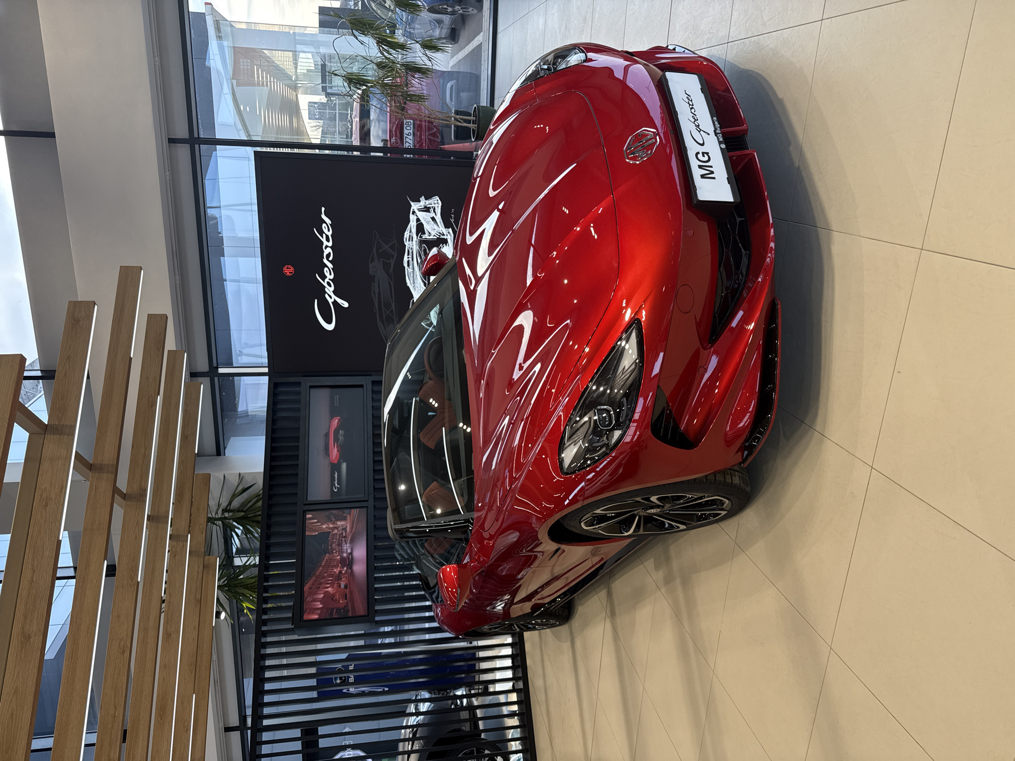 The image displays a bright metallic red MG Cyberster electric roadster prominently positioned on a light-colored tiled floor within a modern automotive showroom. The car faces slightly left, showcasing its angular LED headlights, MG emblem, and a front license plate holder reading "MG Cyberster" and "MG Bulgaria." The vehicle features a black convertible soft top, partially visible brown or tan interior seating, and multi-spoke black and silver alloy wheels.
Behind the car, a black display wall features white "MG Cyberster" branding and a stylized line drawing of the vehicle. Two framed images are mounted on this wall: one depicts a city street at night, and the other shows the rear of a red car. To the left, a vertical slatted dark partition separates the MG display area, with a partial view of a dark grey SUV and a blue pillar marked "Subaru" behind it.
The showroom is illuminated by overhead linear LED light fixtures integrated into a suspended wooden slat ceiling design. Large, floor-to-ceiling glass windows form the exterior wall, allowing natural light and revealing an outdoor parking area. Through these windows, other vehicles are visible, including a red car with a Bulgarian license plate "5776 OB," confirming the location in Sofia, Bulgaria. Potted green plants are placed near the windows. No individuals are present in the scene.