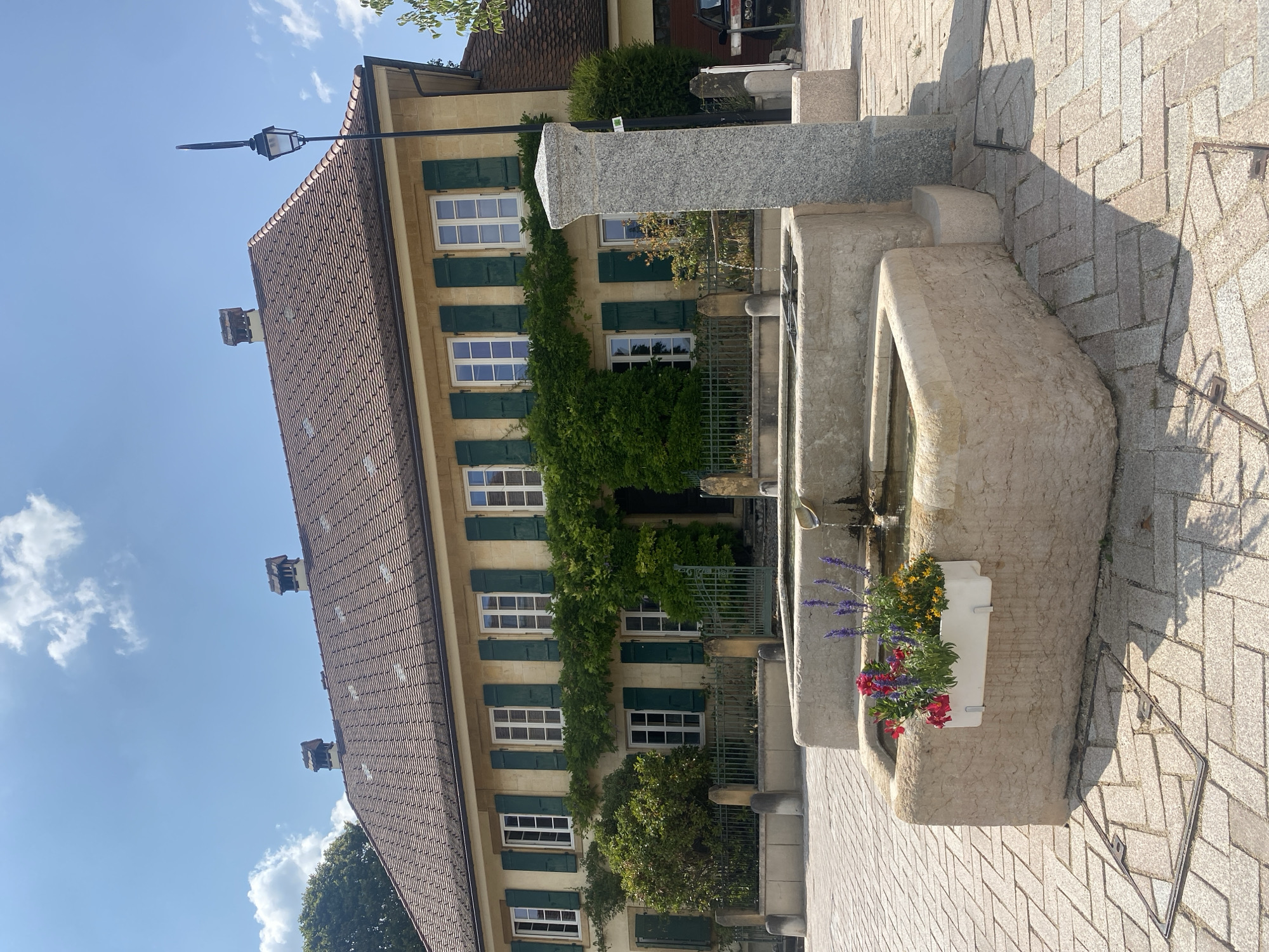 In Milvignes, Switzerland, a multi-tiered stone fountain is centrally located in a paved courtyard. The fountain features a planter box with red and purple flowers. Behind the fountain, a light-yellow building with a red tiled roof stands prominently. The building has multiple windows with closed green shutters and is partially covered by lush green ivy. A black lamppost is visible to the right of the building. The courtyard is paved with light-colored interlocking bricks, and a metal grate is embedded in the pavement in the foreground. The sky is clear with scattered white clouds.