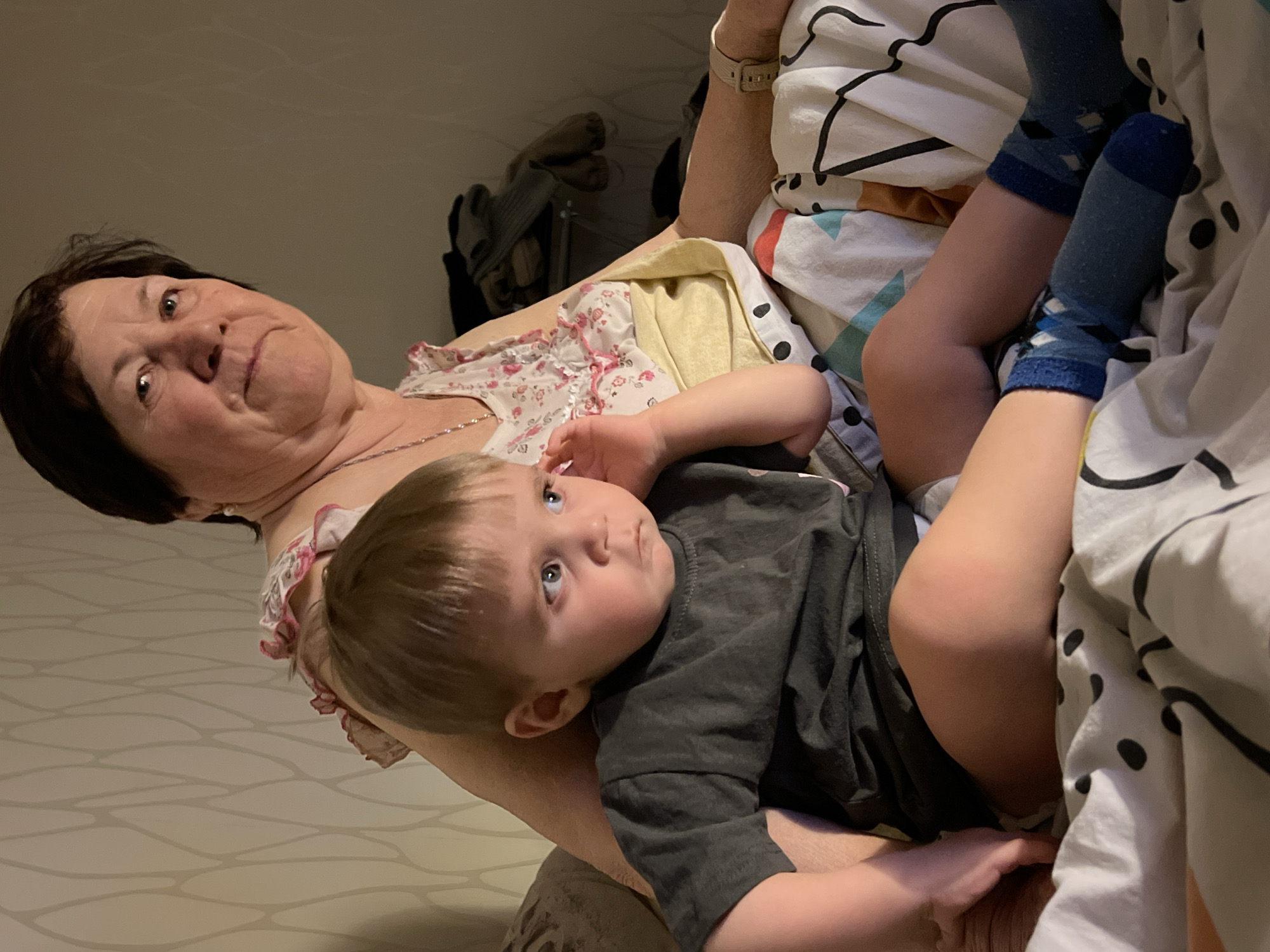 An older woman holds a young child in an indoor setting, likely a home, with soft, subdued lighting. The woman, with dark hair, wears a light-colored top with a pink floral pattern and a delicate necklace, looking towards the camera with a somewhat reserved or observant expression. A watch is visible on her left wrist.
The child, a toddler with light brown hair and bright blue eyes, is dressed in a dark grey t-shirt and blue patterned socks. The child is nestled comfortably against the woman, gazing upwards and to the right, away from the camera, with a calm, curious, or thoughtful expression.
The background includes a patterned duvet or blanket with black lines and dots, suggesting a bed or sofa. Behind them, a light-colored wall features a subtle, wavy pattern. Further in the background, some folded clothes or fabric items are visible, indicating a casual home environment. The scene portrays a quiet, intimate moment between what appears to be a grandmother and grandchild. No text is visible in the image.