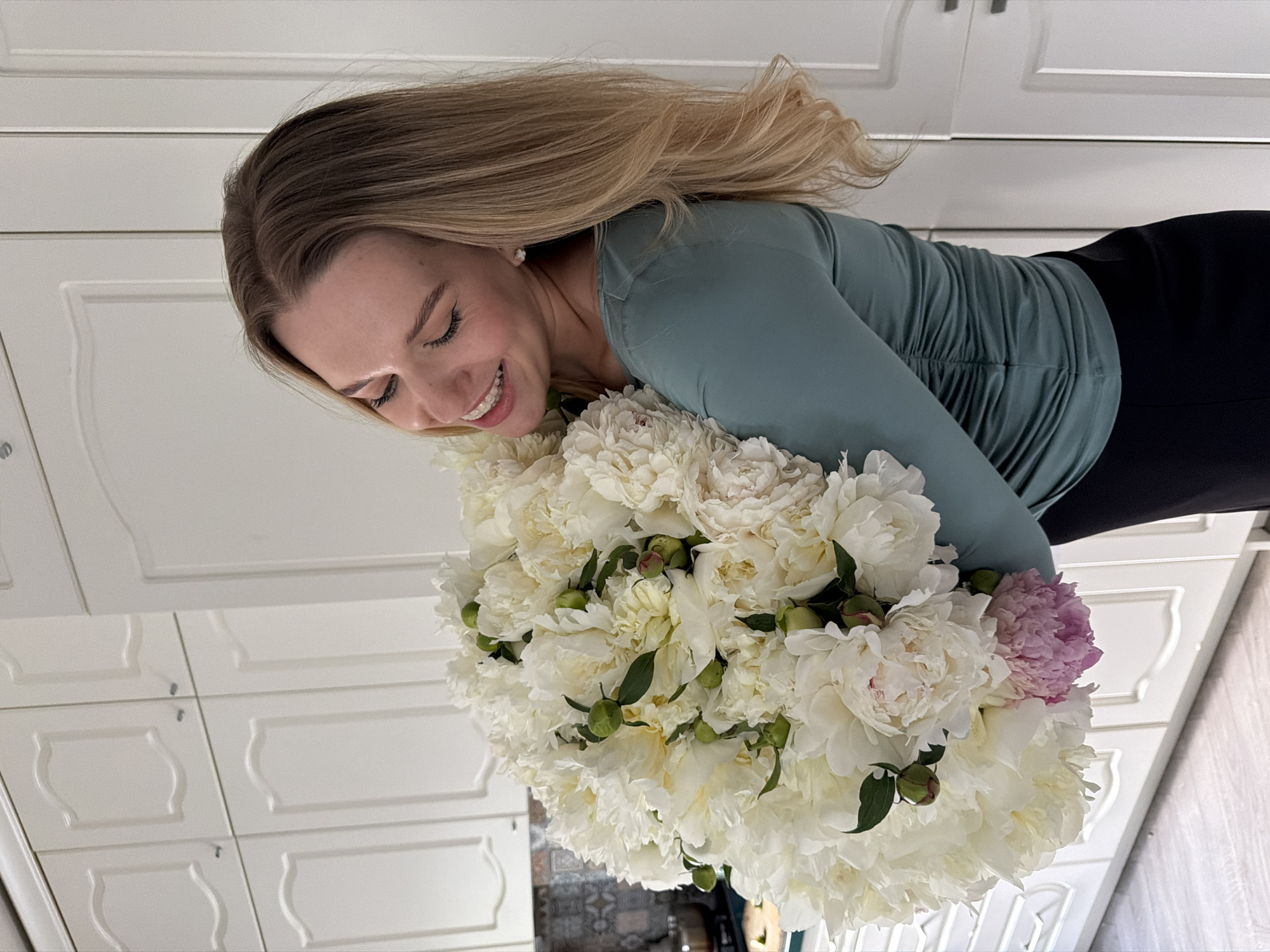 In a residential kitchen with white cabinetry, a young woman with blonde hair embraces a large bouquet of white peonies. She is wearing a teal, ruched long-sleeved shirt and black pants. The woman is smiling and looking down at the flowers. The scene is well-lit, suggesting it was taken indoors during the day.