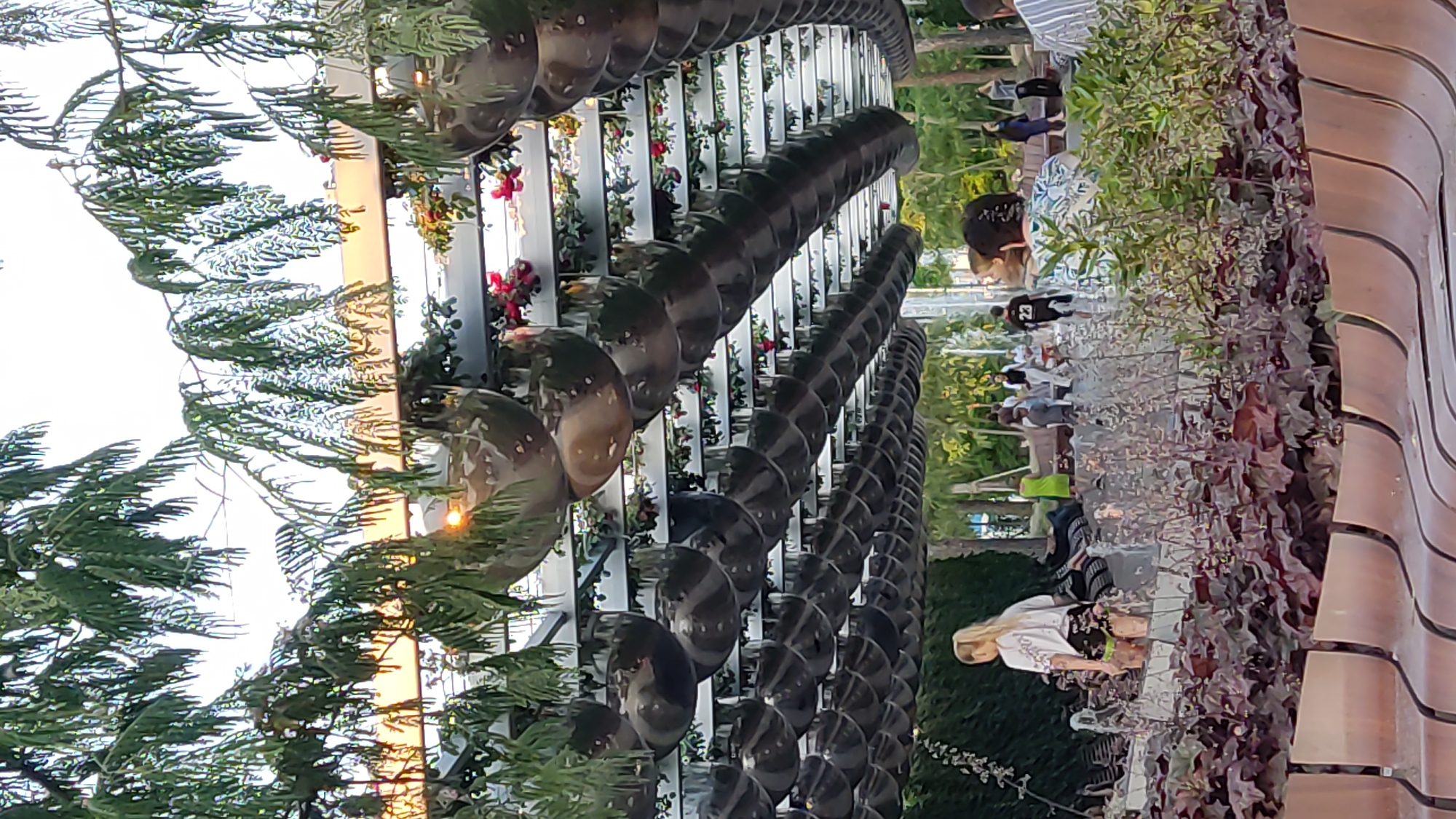 The image depicts an outdoor scene, possibly a park or garden, with a prominent spiral-like metal sculpture. Several people are visible in the background, some walking and others seated. A woman with blonde hair is sitting on a bench, looking towards the sculpture. The setting appears to be a public space, with lush greenery and flowers adorning the structure. The time of day is indicated by the warm, soft light, suggesting it might be late afternoon or early evening. The overall atmosphere seems relaxed and leisurely. The location is identified as Krasnodar, Russia.