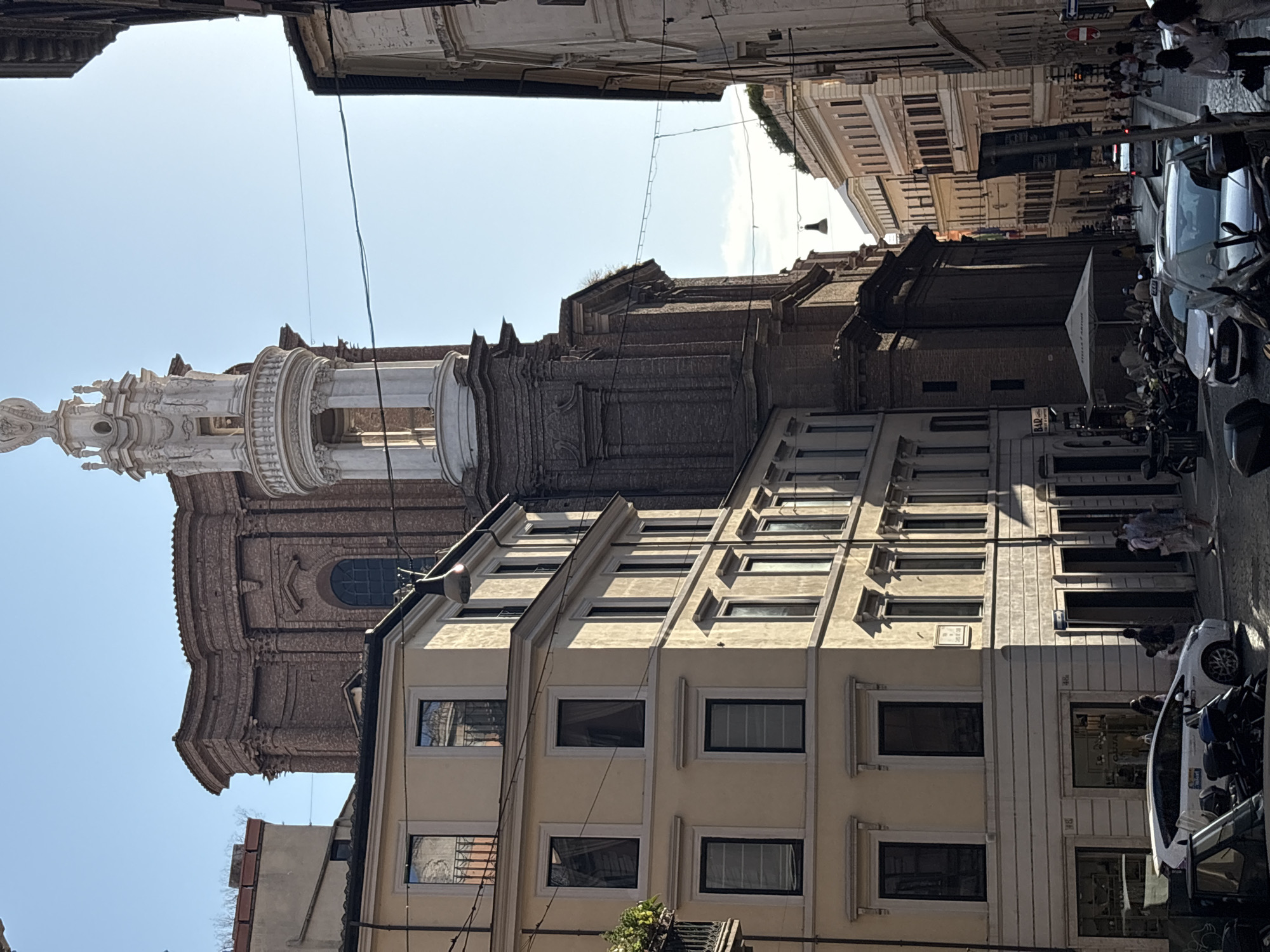 This is a street view in Rome, Italy, under a clear blue sky. The scene is dominated by the architecture of buildings and a prominent church tower. On the left, a tall, multi-story building with cream-colored facade and numerous dark-framed windows is illuminated by sunlight, casting shadows. A white car is parked in front of its ground-level shops. Adjacent to it, a larger, dark brick structure, part of a church, rises higher, featuring ornate brickwork and a distinctive white tower with multiple levels and architectural embellishments. The tower is topped with a decorative element and has visible columns and open spaces.
The street itself is paved with cobblestones. Several people are visible walking on the sidewalk, including a group on the right side of the street. Motorcycles and scooters are parked along the side of the street, and a few cars are present, including a white taxi. On the right side of the frame, another building with a lighter facade is visible, showing a similar architectural style with many windows. Power lines are strung across the street, with a hanging light fixture visible above. Further down the street, the buildings recede, creating a sense of depth, with more people and vehicles present in the distance. The overall impression is of a bustling urban environment in a historic European city.