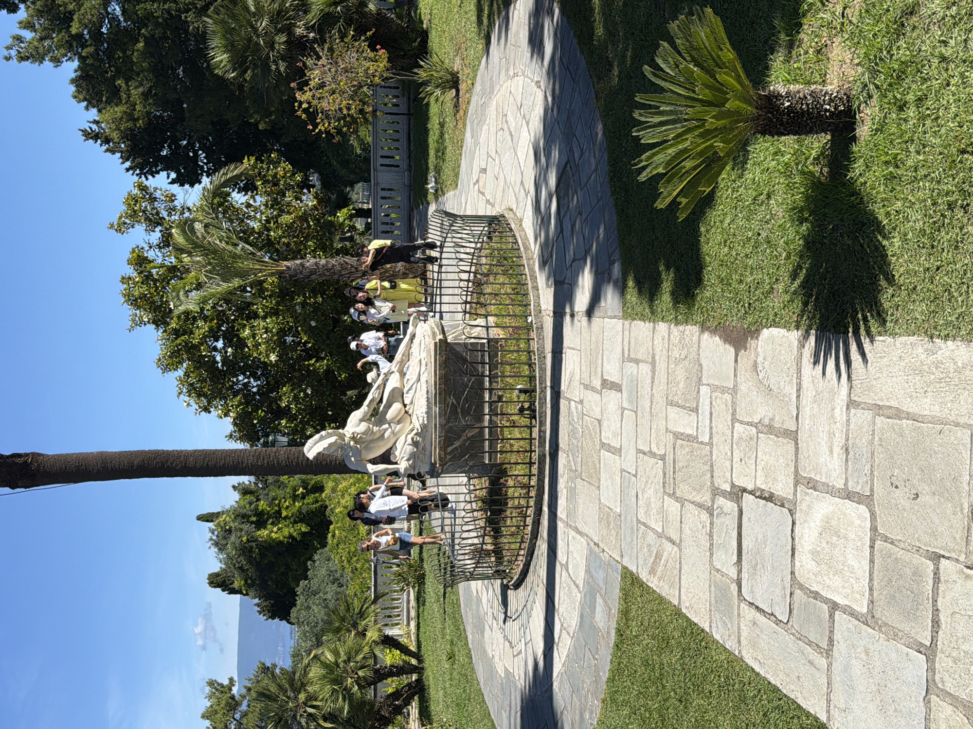 The image presents a sunny outdoor scene, likely a garden or park in Gastouri, Greece. A wide, curved pathway, paved with irregular light-colored stone blocks, leads through well-maintained green grass on the right side of the frame. Bright sunlight casts strong, defined shadows of trees and a low metal fence across the pathway and lawn, indicating a clear, sunny day, possibly mid-morning to afternoon.
In the mid-ground, a large white classical-style statue of a reclining figure is a central focal point. It is enclosed by a semicircular black metal fence. Several people are gathered around the statue, observing it or taking photos. They are casually dressed, with some individuals clearly visible, such as a person in a yellow top and others in white and dark clothing.
The setting is rich with lush vegetation, including tall palm trees with visible trunks and vibrant green fronds, and dense green foliage from various other trees and shrubs. The upper left portion of the image shows a canopy of dark green trees against a bright, clear blue sky. In the background, beyond the immediate garden, distant hazy mountains or hills are visible under the expansive blue sky, suggesting a scenic overlook, possibly towards a body of water or another landmass. There is no visible text in the image.