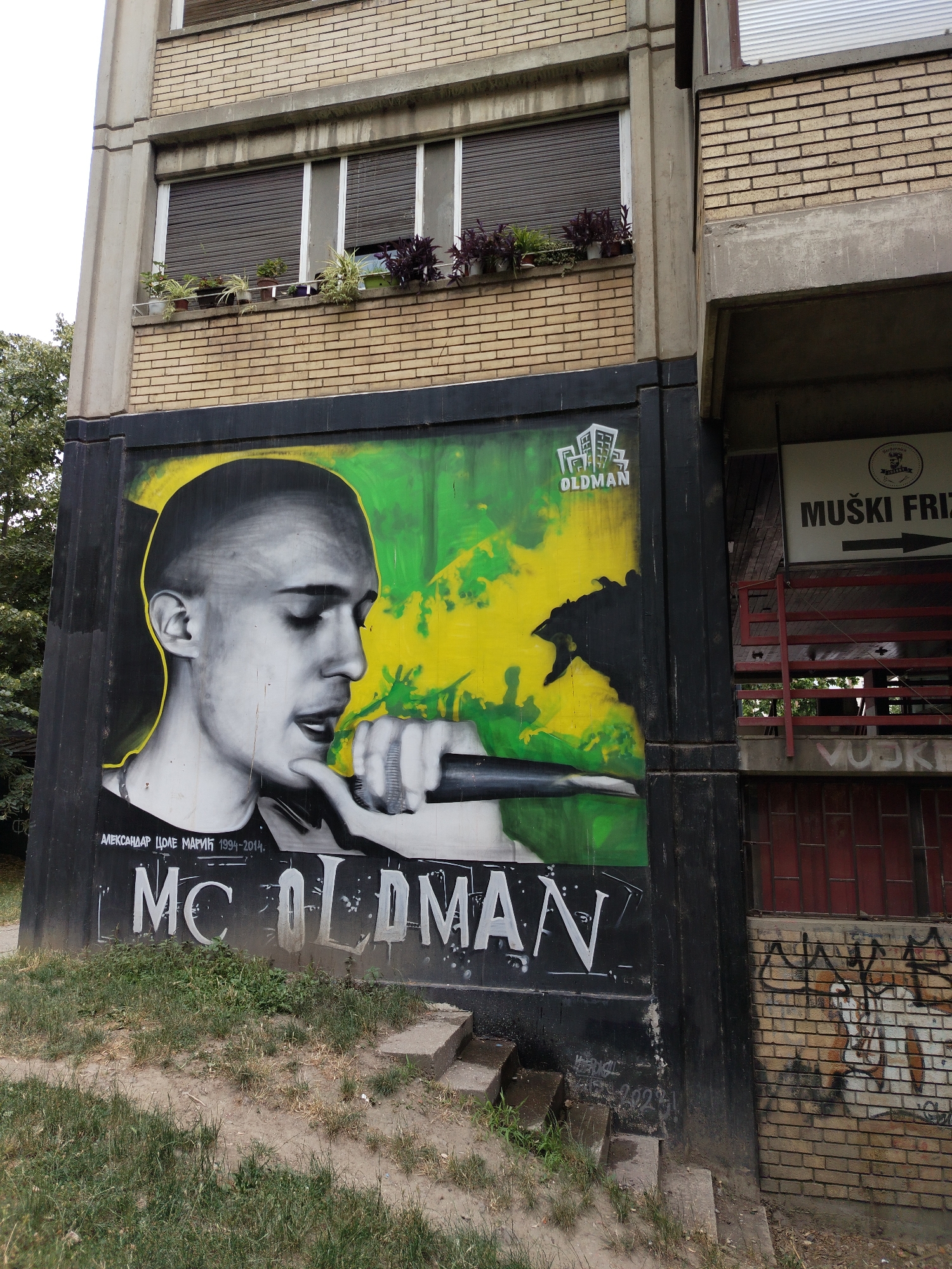 The image shows a mural of a man, identified as "MC OLDMAN," on the side of a building. The man is depicted in black and white, with a yellow outline, and is holding a microphone to his mouth. The mural is placed next to an entrance to a barber shop, indicated by a sign that reads "MUŠKI FRIZ" (men's hair salon). The building appears to be old, with a brick facade and metal shutters on the windows. Plants are visible on a ledge above the mural. The overall scene suggests an urban environment, likely in Novi Sad, Serbia, given the Cyrillic lettering on the barber shop sign. There is no other visible human presence in the image.