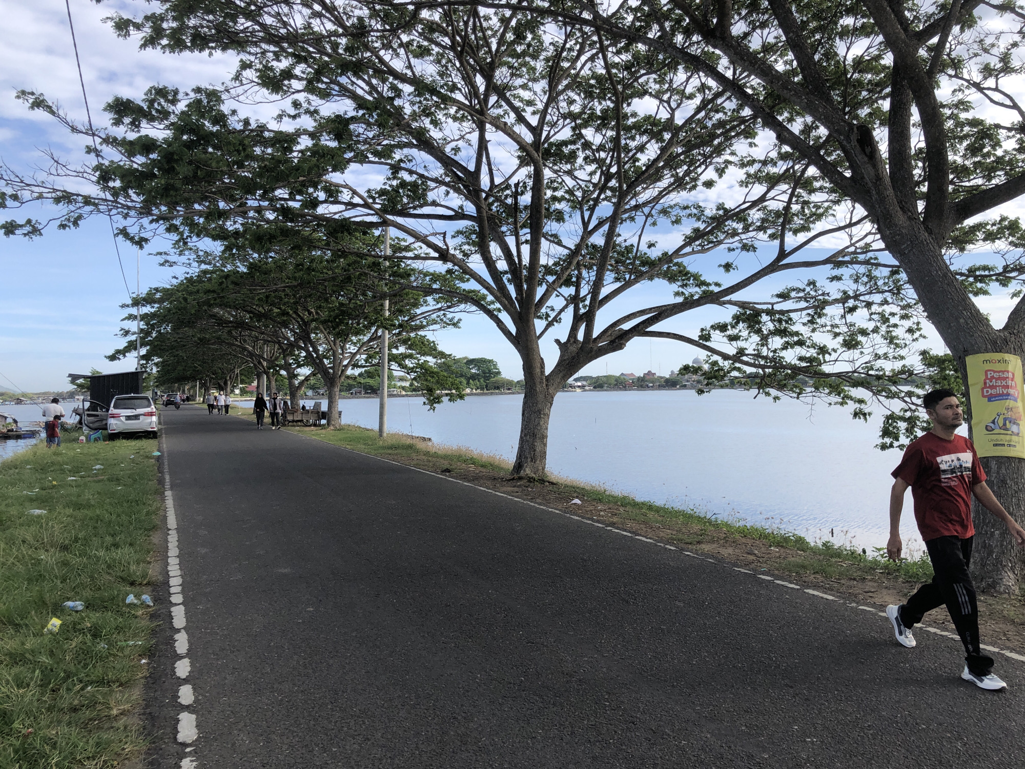 The image captures a scene along a road bordering a lake in Lhokseumawe, Indonesia. A man walks along the right side of the road, heading away from the camera. He wears a maroon t-shirt, black pants with white stripes, and white sneakers. A sign is posted on a tree next to him, partially obscured.
On the left side of the road, there is a grassy area with scattered debris and trash. A white car is parked near the edge of the road. Further down, several people are gathered near the lake's edge, with one person standing by the water.
The road itself is asphalt and shows signs of wear. Trees line both sides of the road, creating a canopy overhead. The lake is calm, reflecting the sky, and some buildings are visible on the far shore. The sky is blue with scattered clouds.