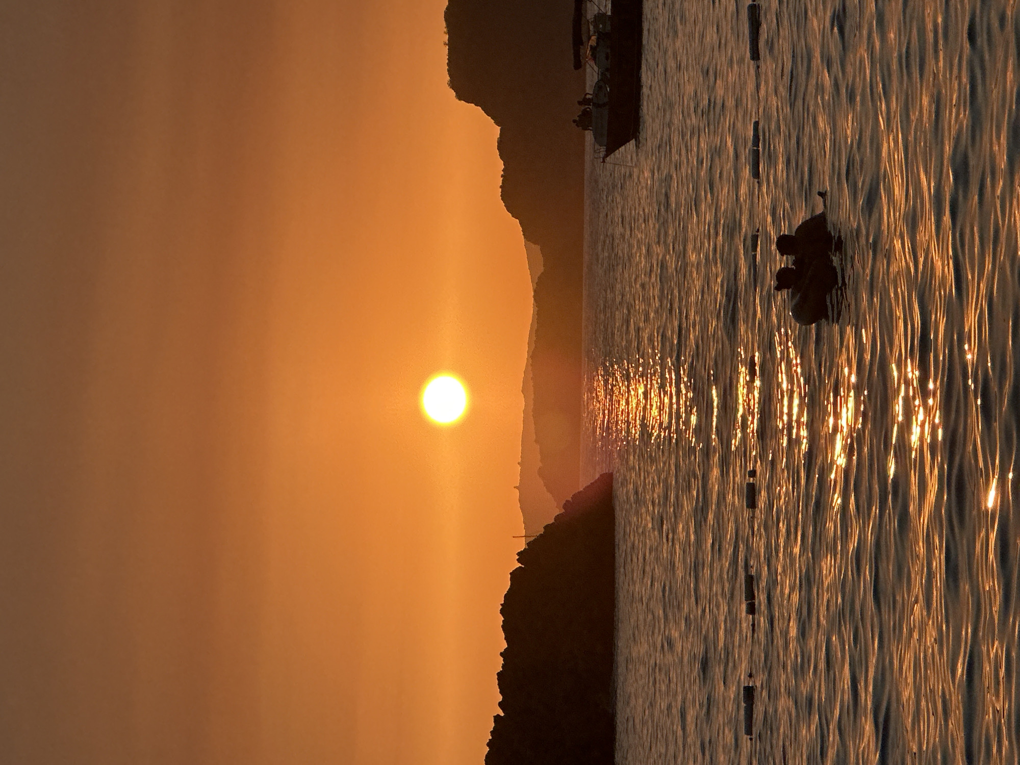 A sunset scene unfolds over a body of water, likely the Adriatic Sea near Sveti Stefan, Montenegro. The sun, a bright orb, hangs low in the sky, casting a warm, orange glow that saturates the entire image. The sky transitions from a lighter orange near the horizon to a deeper, more muted orange at the top of the frame.
The water, choppy with visible ripples, reflects the sunset's light, creating a shimmering path across its surface. In the foreground, two figures are partially submerged in the water, seated in what appears to be a small, inflatable boat or raft. They are silhouetted against the bright water, their forms indistinct.
Further back, on the left side of the frame, a rocky, tree-covered island juts out of the water, silhouetted by the sunset. On the right side, another silhouetted landmass, likely a hillside or cliff face, is visible. A boat, also in silhouette, is moored on the right side of the water, with several figures standing on its deck. In the distance, faint outlines of hills or mountains are visible, adding depth to the landscape. A series of rectangular buoys or markers are partially visible in the water, extending horizontally across the frame.