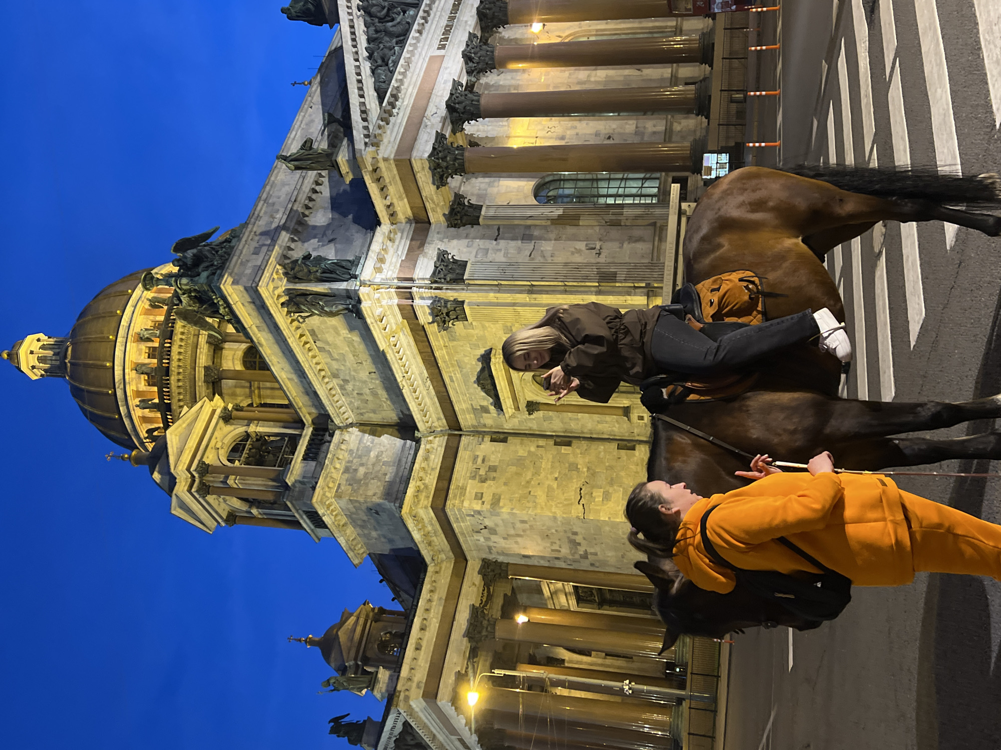 Two women are riding horses on a street in Saint Petersburg, Russia, at night. They are positioned on a crosswalk in front of Saint Isaac's Cathedral. One woman is sitting on the horse, and the other is walking alongside it. The woman on the horse is wearing a brown jacket, black jeans, and white shoes. The other woman is wearing an orange tracksuit and a black backpack. The street is illuminated by streetlights. The sky is dark blue.