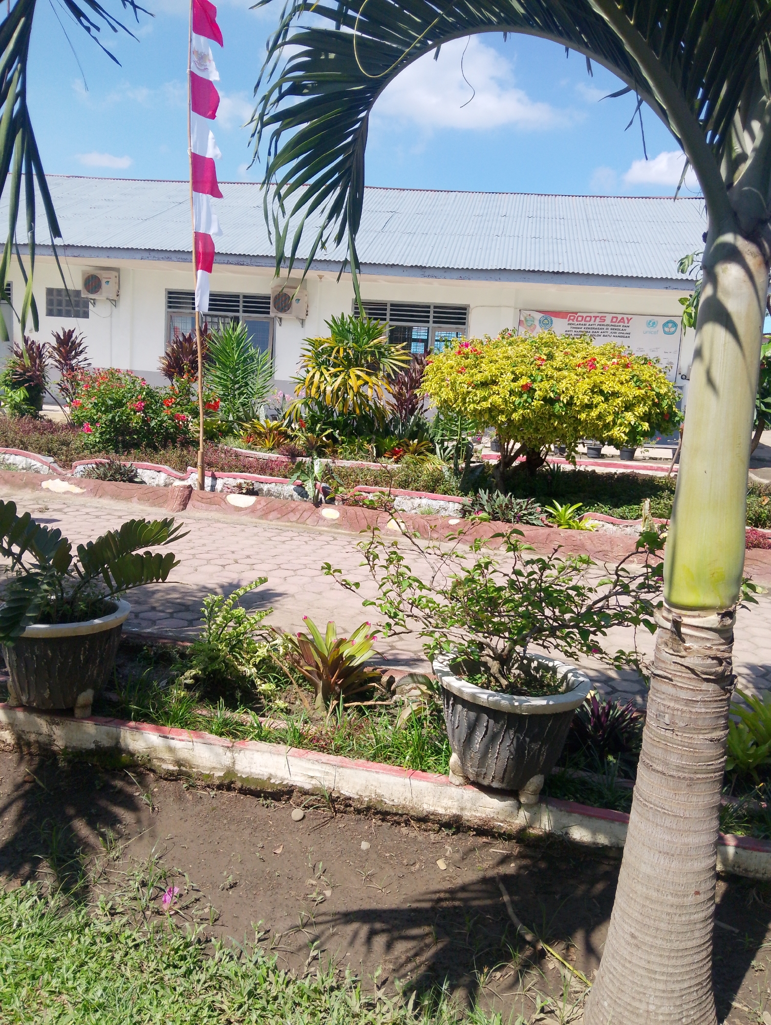 A low-angle, outdoor shot shows a palm tree trunk on the right, with a portion of its fronds arching over the top. The scene appears to be in the city of Serbelawan, Indonesia. In the background, a white building with a corrugated metal roof stands under a bright blue sky with some scattered clouds. Air conditioning units are visible on the building's exterior. A flagpole with a red and white Indonesian flag is positioned in front of the building, partially obscured by palm fronds. The foreground features a paved walkway with a decorative border and a garden bed filled with various plants, including potted greenery. The garden bed is bordered by a low wall. Shadows of palm fronds are cast across the ground. A banner with text, including "ROOTS DAY," is visible on the side of the building.