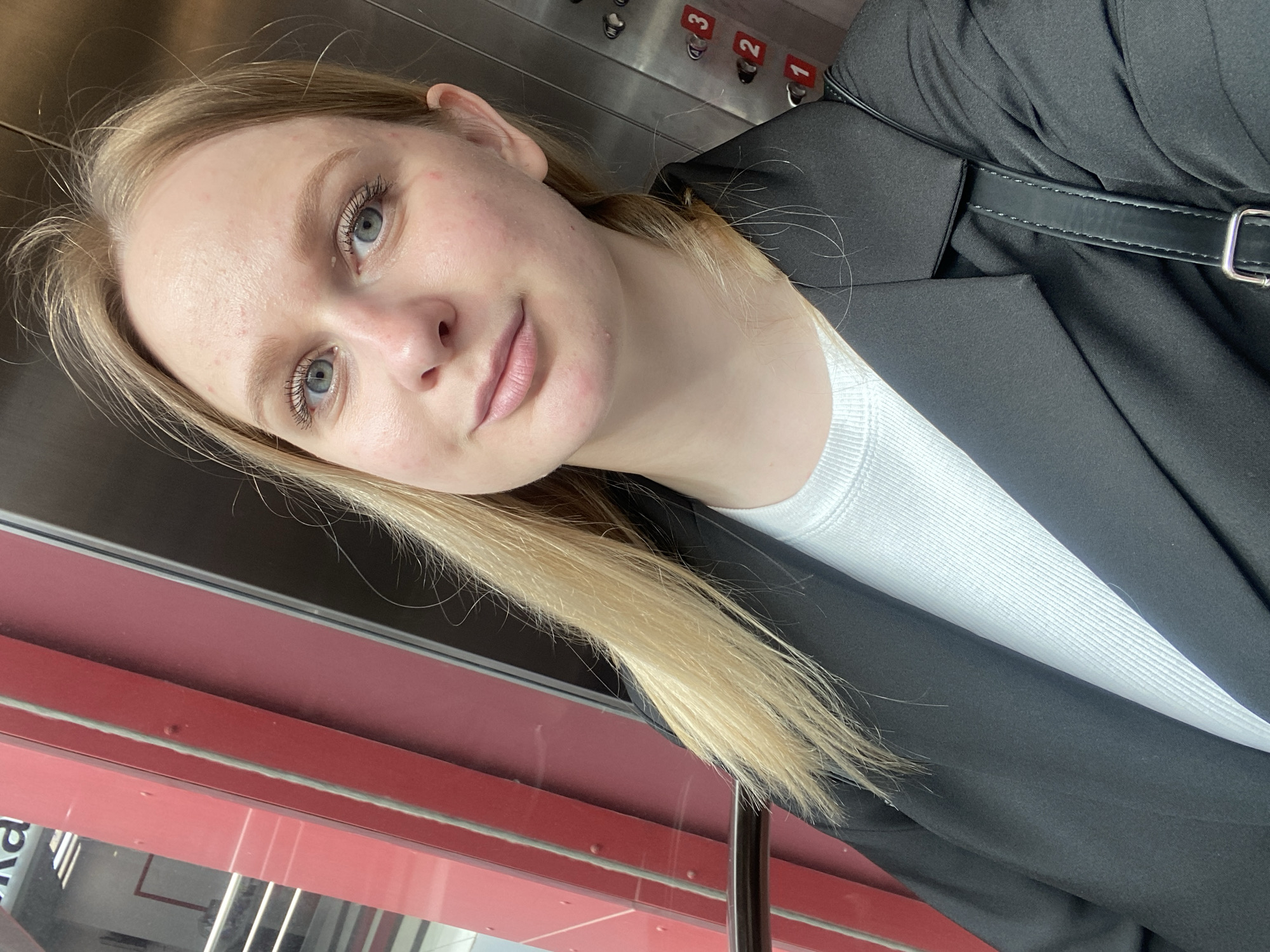 The media file is a portrait of a young woman with blonde hair and blue eyes, taken in an elevator. She is wearing a black blazer over a white ribbed top. Her hair is down and falls over her shoulder. The elevator panel is visible in the background with buttons numbered 1, 2, and 3. There is a red panel on the left side of the image, possibly part of the elevator's interior or the building's structure. The lighting suggests it is daytime. The woman's expression is neutral, and she appears to be looking at the camera. The scene is indoors, within an elevator, likely in Izhevsk, Russia, given the location context.
