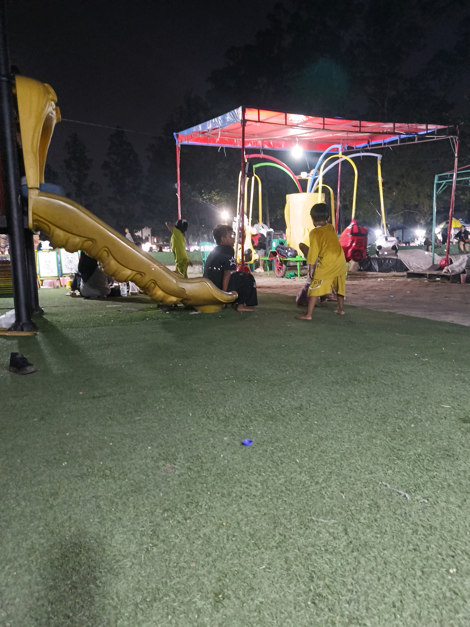 The image depicts a nighttime scene at a playground in Tangerang, Indonesia. A large yellow slide is a prominent feature on the left side of the frame. In the center, two young children are present: one wearing a black shirt and sitting, and another in a yellow shirt standing. Behind them, a red and blue canopy covers a structure with yellow poles, possibly part of a swing set or a carnival ride. Several other figures are visible in the background, along with tents and lights, suggesting a lively evening atmosphere. The ground is covered in green artificial turf.