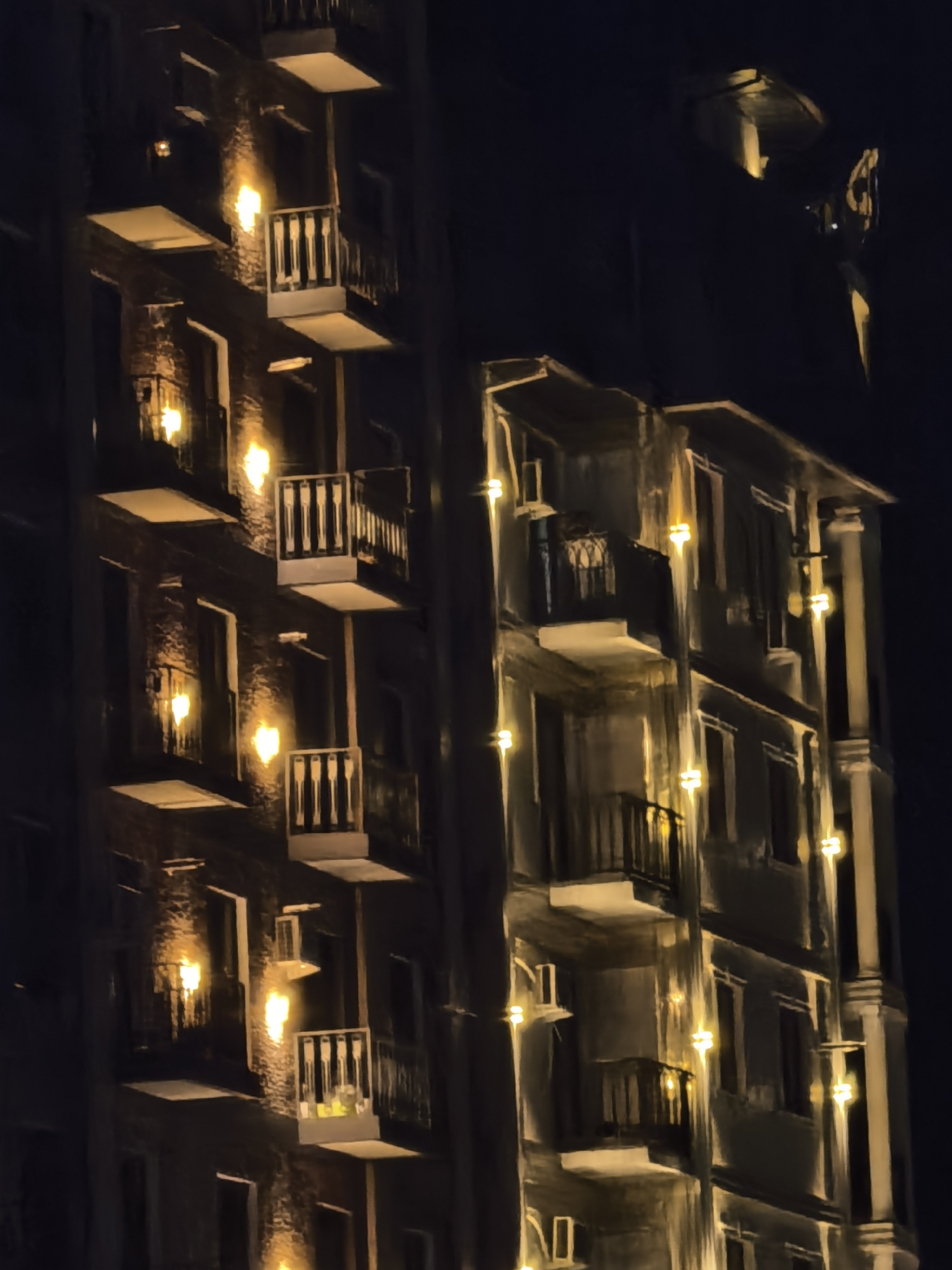 The image depicts the facade of an apartment building at night in Fergana City, Uzbekistan. The building has multiple balconies with railings, some of which are illuminated by warm, yellow lights. The architecture appears to be a mix of brickwork and plastered surfaces. The lighting suggests that some of the apartments are occupied. The overall scene is dark, with the building's lights providing the primary illumination against the night sky.