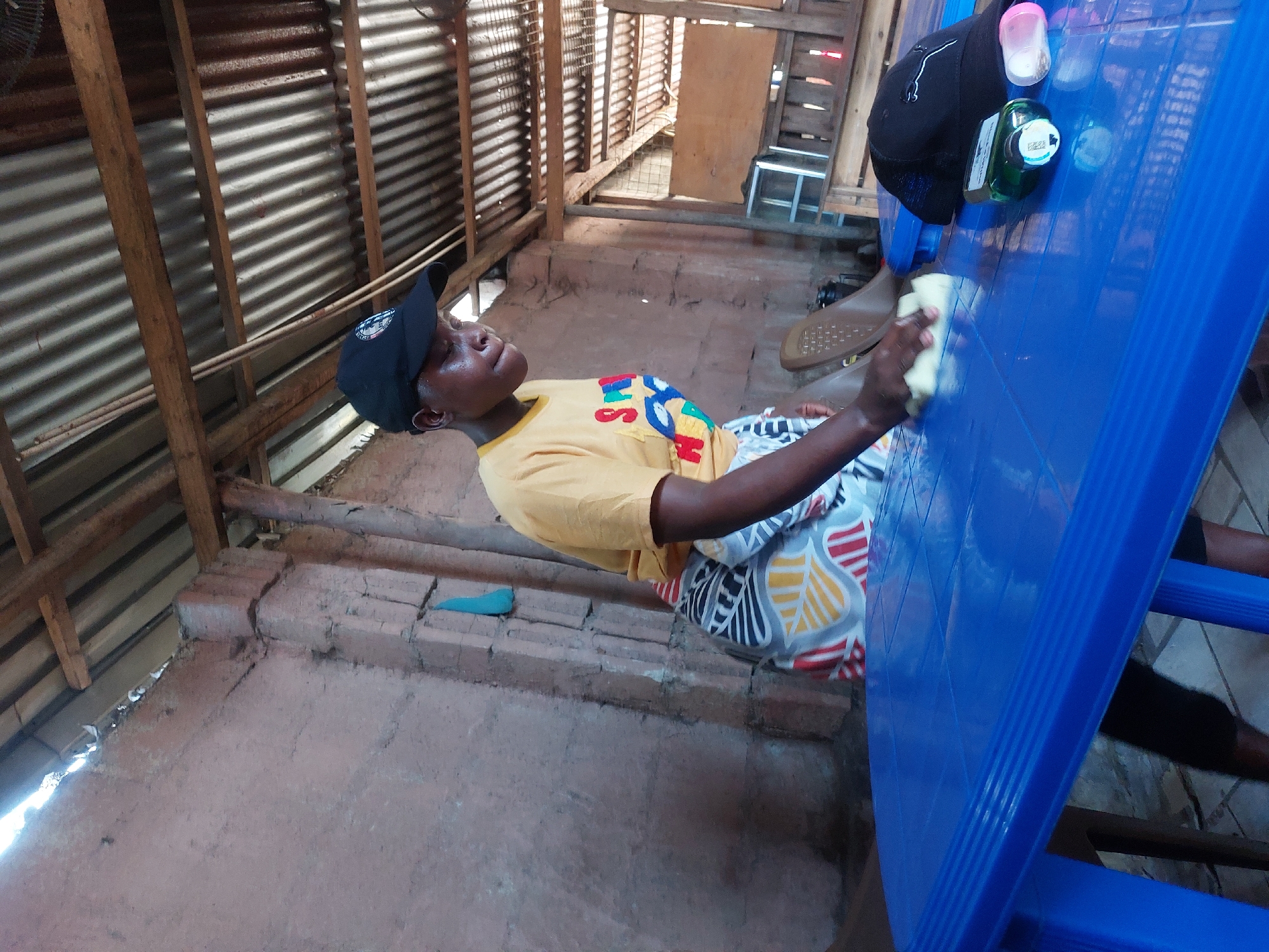 An indoor scene in Dar es-Salaam, Tanzania, depicts an individual engaged in cleaning. A dark-skinned female, approximately in her twenties, stands on the left side of a bright blue rectangular plastic table. She wears a yellow short-sleeved t-shirt with a multi-colored graphic print, a black baseball cap, and a patterned wrap-around skirt over dark trousers. Her right hand holds a light-colored sponge or cloth, which she is actively using to wipe the table surface. Her gaze is directed downwards at the table.
On the right side of the table, a black baseball cap (with a white emblem), a green bottle with a white cap (contents unknown, possibly cleaning agent), and a small pink plastic container are visible. The immediate background consists of rough, reddish-brown plastered walls, exhibiting visible cracks and uneven texture. A section of a brick support column is present, from which a light blue piece of fabric hangs. The ceiling is constructed from corrugated metal sheeting supported by exposed wooden beams and rafters. Gaps are visible between the top of the wall and the roofline, allowing natural light to enter. Further in the background, a light brown plastic chair is partially visible, along with structures made of wooden slats and wire mesh.