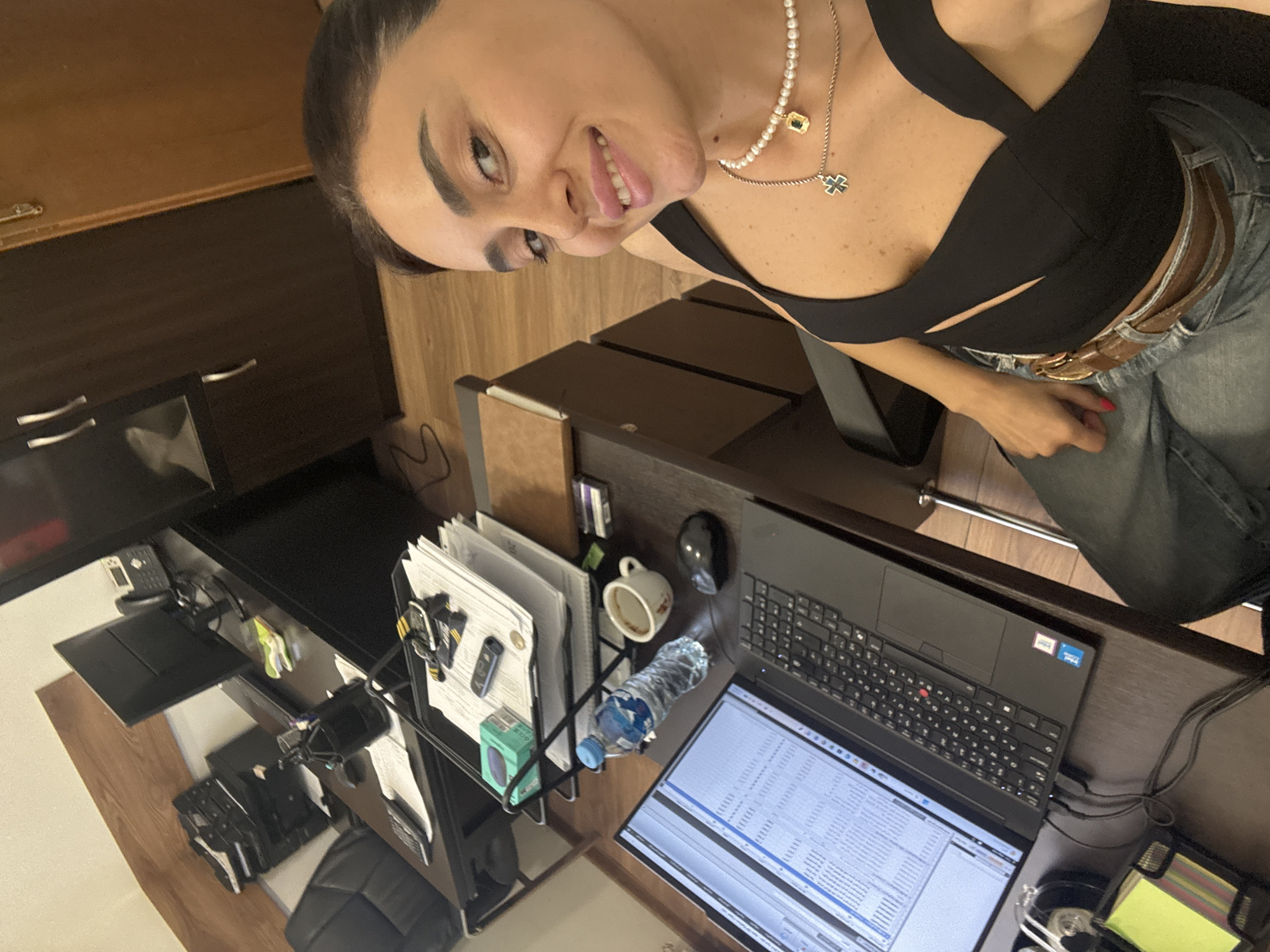A medium shot photo shows a woman with dark hair and brown eyes in a black top and blue jeans. She is sitting at a desk in what appears to be an office. The desk is cluttered with a laptop, a bottle of water, a coffee mug, a mouse, and papers. Behind the desk are cabinets and shelves. The woman is smiling and looking at the camera. The scene is well-lit and the colors are natural.