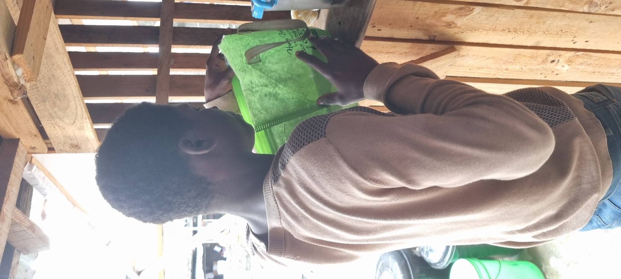 A medium shot from behind shows a young Black person wearing a tan crewneck sweater and blue jeans. The person's right hand is visible near a green reusable bag, and their left hand is holding the bag open. The person is standing in what appears to be an outdoor market stall constructed with wooden planks. In the background, a blue container is visible on a shelf. The image is set in Lagos, Nigeria.