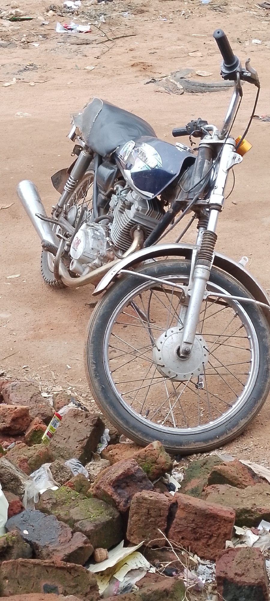 An abandoned motorcycle rests on a patch of sandy, dirt ground, surrounded by scattered debris. The motorcycle, a dark blue and silver color, appears to be in a state of disrepair. Its seat is torn, and some parts of the bodywork are damaged. The engine, exhaust pipe, and wheels are visible, suggesting it is a relatively small displacement motorcycle. The ground is littered with various items, including broken bricks, plastic bags, and other unidentifiable trash, indicating an urban or semi-urban environment. The overall impression is one of neglect and abandonment in a somewhat neglected outdoor space.