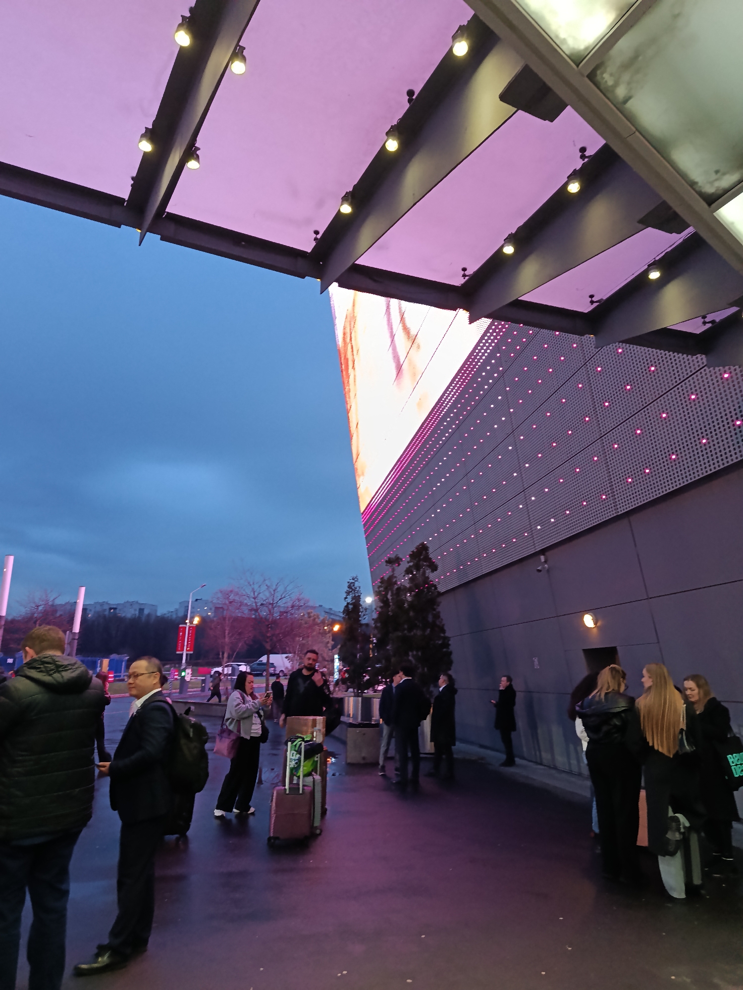 The image depicts an evening scene outside what appears to be a modern building with a large LED screen and a covered entrance in Khimki, Russia. A group of people are standing and conversing near the building's entrance. Some people have luggage, suggesting they may be travelers or visitors to the location. The sky is overcast, and the artificial lighting from the building and surrounding street lamps illuminates the area.