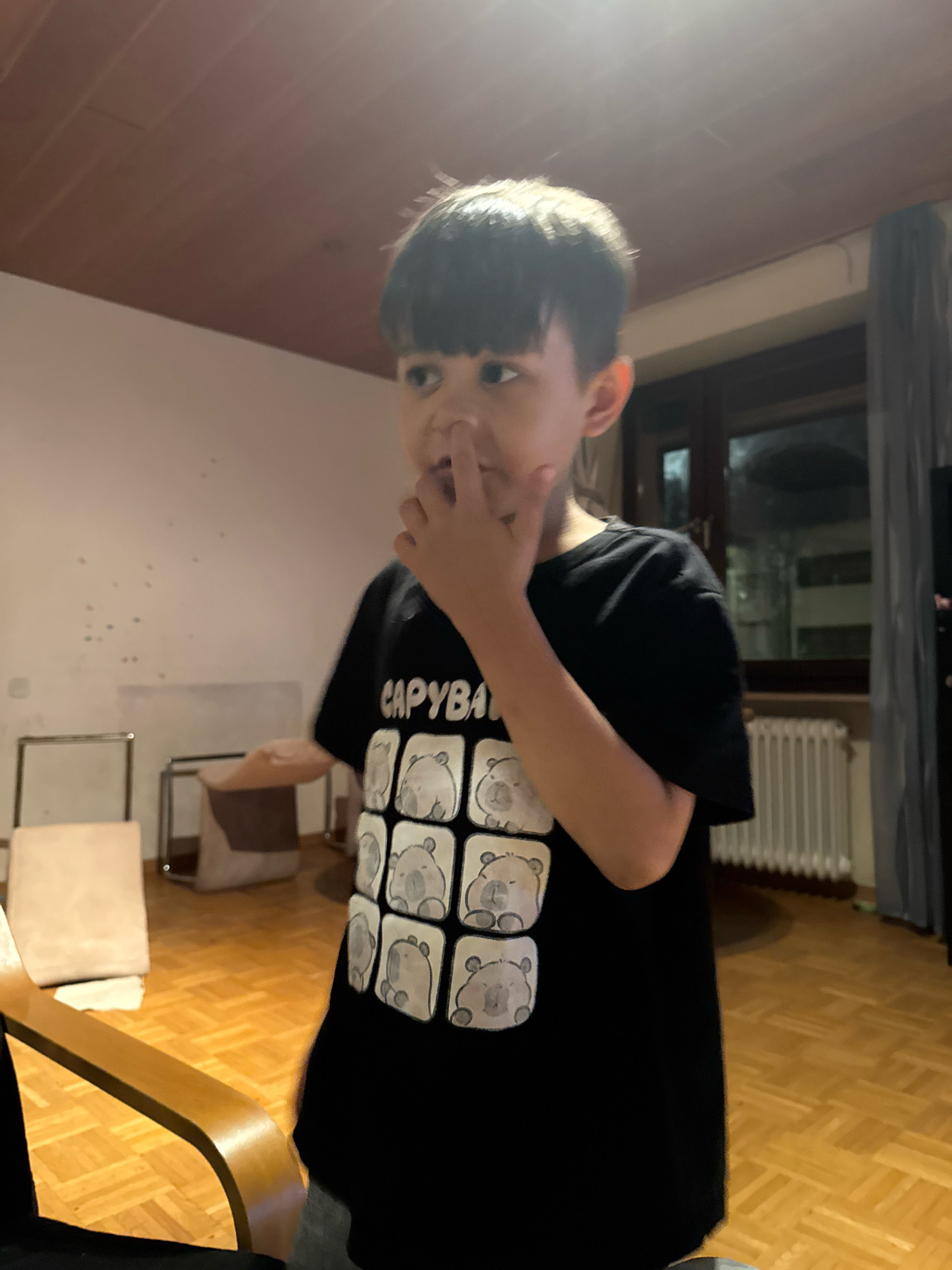 A young boy is standing indoors, looking to his left with his right index finger touching his nose. He is wearing a black t-shirt with a repeating pattern of cartoon bears and the word "CAPYBARA" printed above them. The room features wooden flooring, a white wall with some scattered colorful dots, a radiator under a window with gray curtains, and furniture including chairs and a small table. The overall lighting suggests it is evening or the room is dimly lit. The image appears to be taken in a residential setting, potentially a living room or playroom. There are no visible indications of the boy's exact location within Überherrn, Germany.