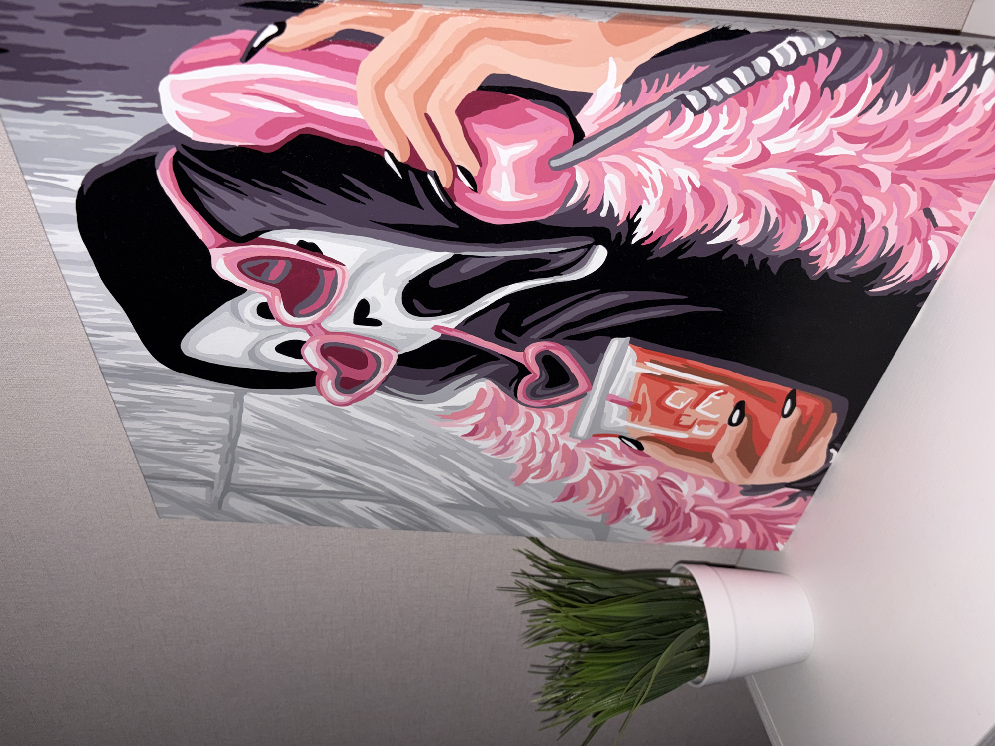The image shows a print hanging on a wall. The print depicts a stylized scene with a woman with dark hair, heart-shaped sunglasses, and a pink feather boa. She is holding a pink object with long, manicured nails. The print also includes an object that resembles a soda can. Below the print, there is a small potted plant with green foliage. The background appears to be an interior setting, possibly a room with light-colored walls. Based on the information provided, the location is Saratovskaya, Russia.