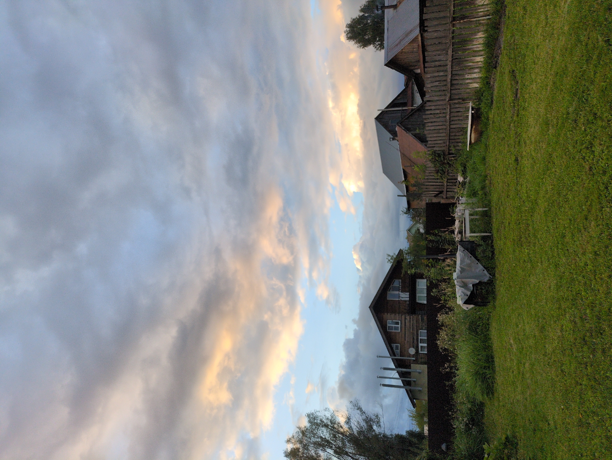The image depicts a rural or suburban scene in Kulebaki, Russia, likely during dusk, as indicated by the dramatic sky filled with clouds and streaks of warm, sunset light. In the foreground, a lush green lawn stretches towards a wooden fence. Beyond the fence, several rustic wooden houses are visible, some with corrugated metal roofs. One house, larger than the others, is made of dark wood and has multiple windows and chimneys. There are also various objects scattered around the yard, including a white table and an old bathtub placed on the grass near the fence. The overall atmosphere is serene and slightly melancholic, characteristic of a quiet evening in the countryside.