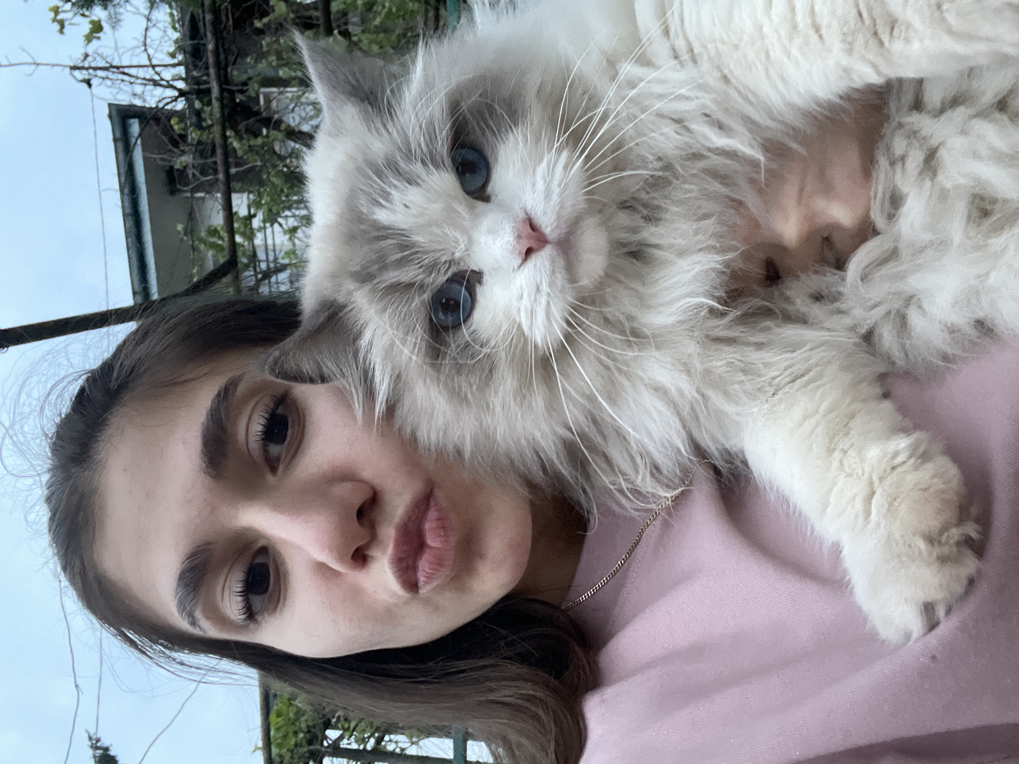 A young woman is holding a fluffy, long-haired cat with blue eyes close to her face. The woman has dark hair and is wearing a pink top. The cat has a predominantly white coat with grey markings on its face and ears. They appear to be outdoors, with foliage and a building visible in the background. The image's orientation is vertical. The exact location in Sofia, Bulgaria, cannot be determined from the image alone.