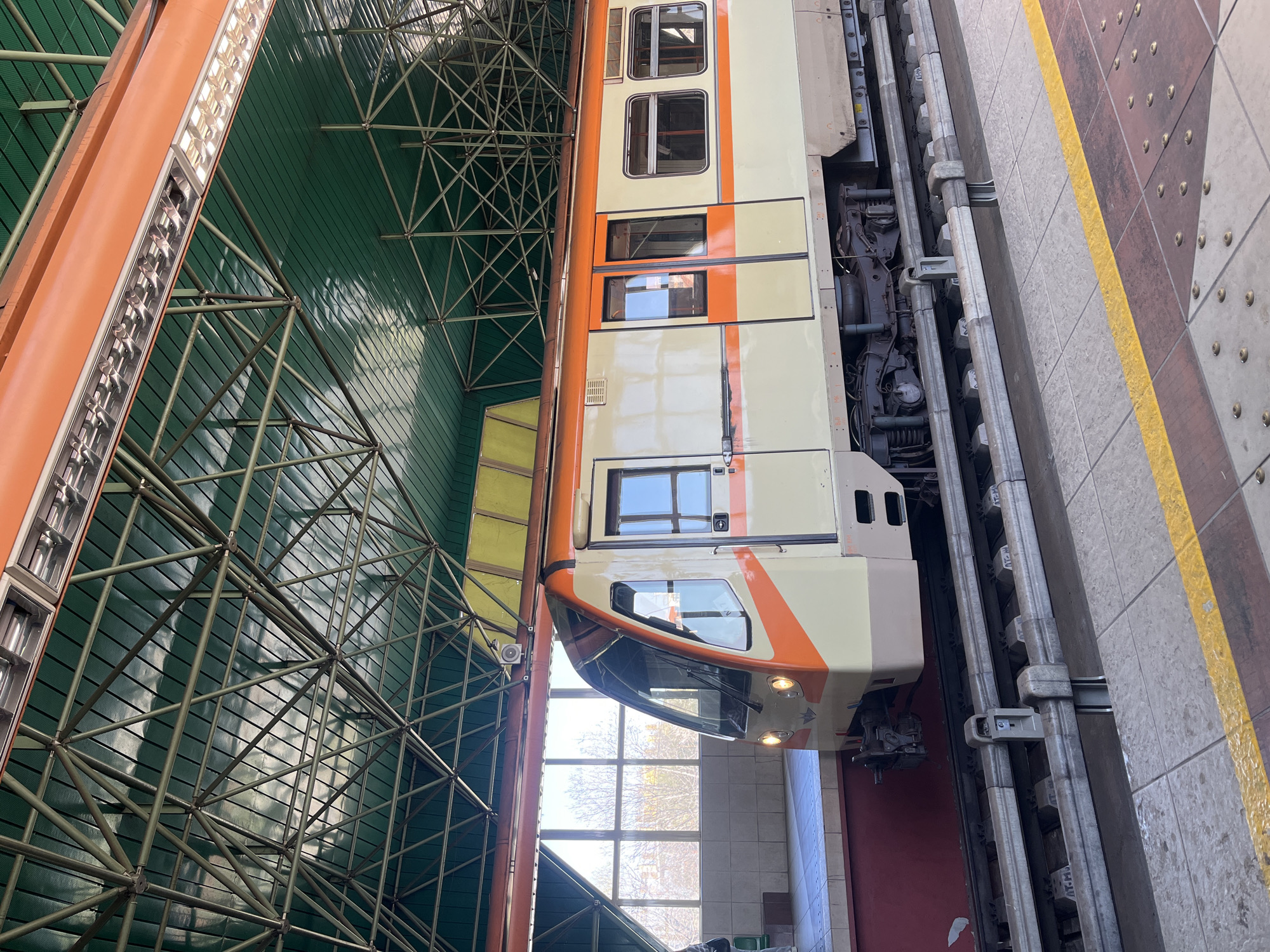 The image shows a train at a station in Sofia, Bulgaria. The train is predominantly cream and orange, with a clear windshield and illuminated headlights. It is parked next to a platform with a yellow safety line. The station's interior features a green-colored ceiling supported by a complex network of metal beams. In the background, a window provides a view of the outside. The train appears to be ready for departure or arrival at the station.