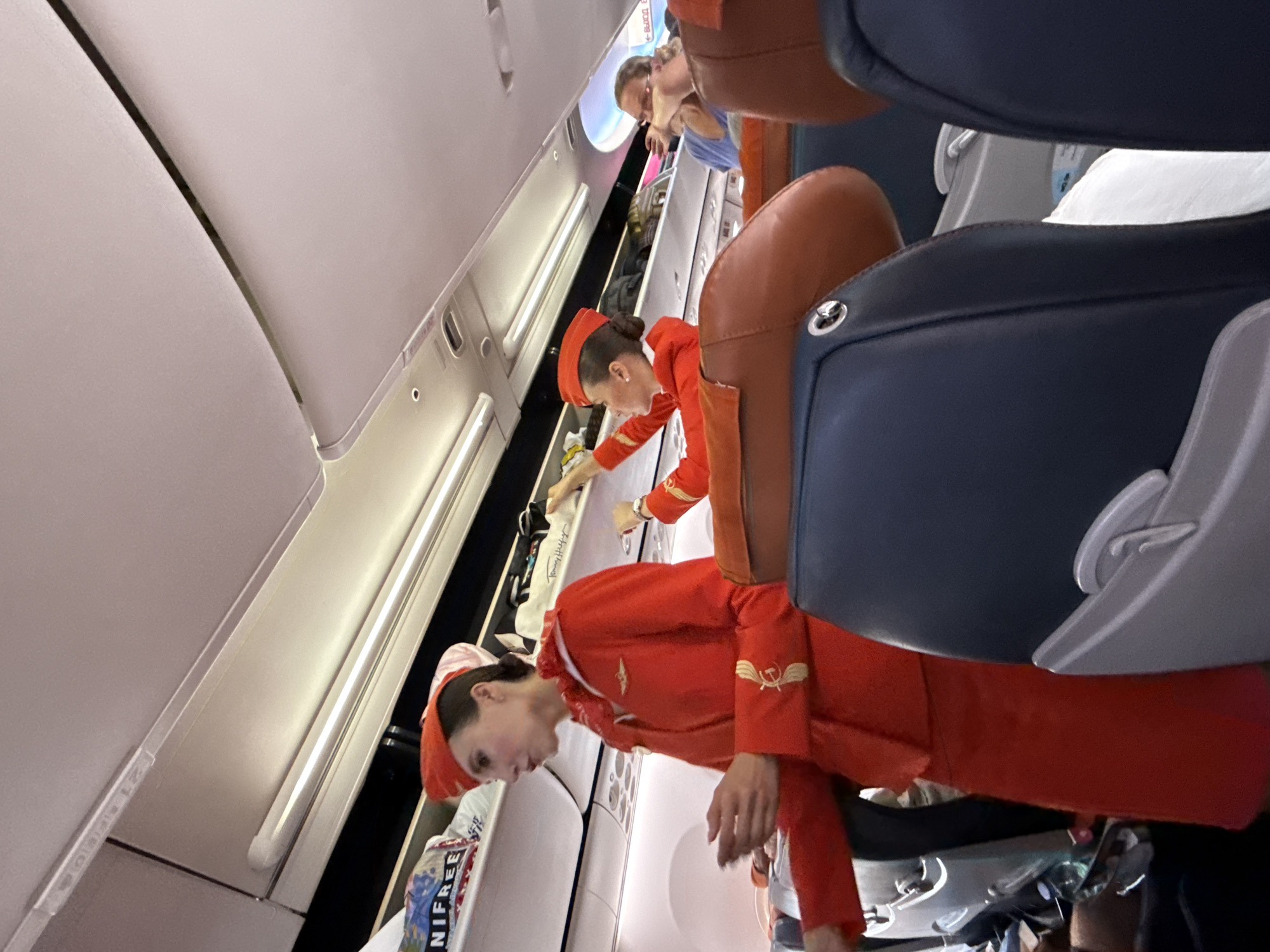 The image shows the interior of an airplane cabin. Two flight attendants in red uniforms are visible. The flight attendant on the left is standing and looking forward, with her right hand gesturing. The flight attendant on the right is reaching into an overhead compartment. Several passengers are seated in the foreground, with their heads and shoulders visible. Overhead compartments are filled with luggage. The lighting suggests it is daytime. This scene is occurring inside an airplane, not a specific location within the city of Arnavutköy, Turkey.