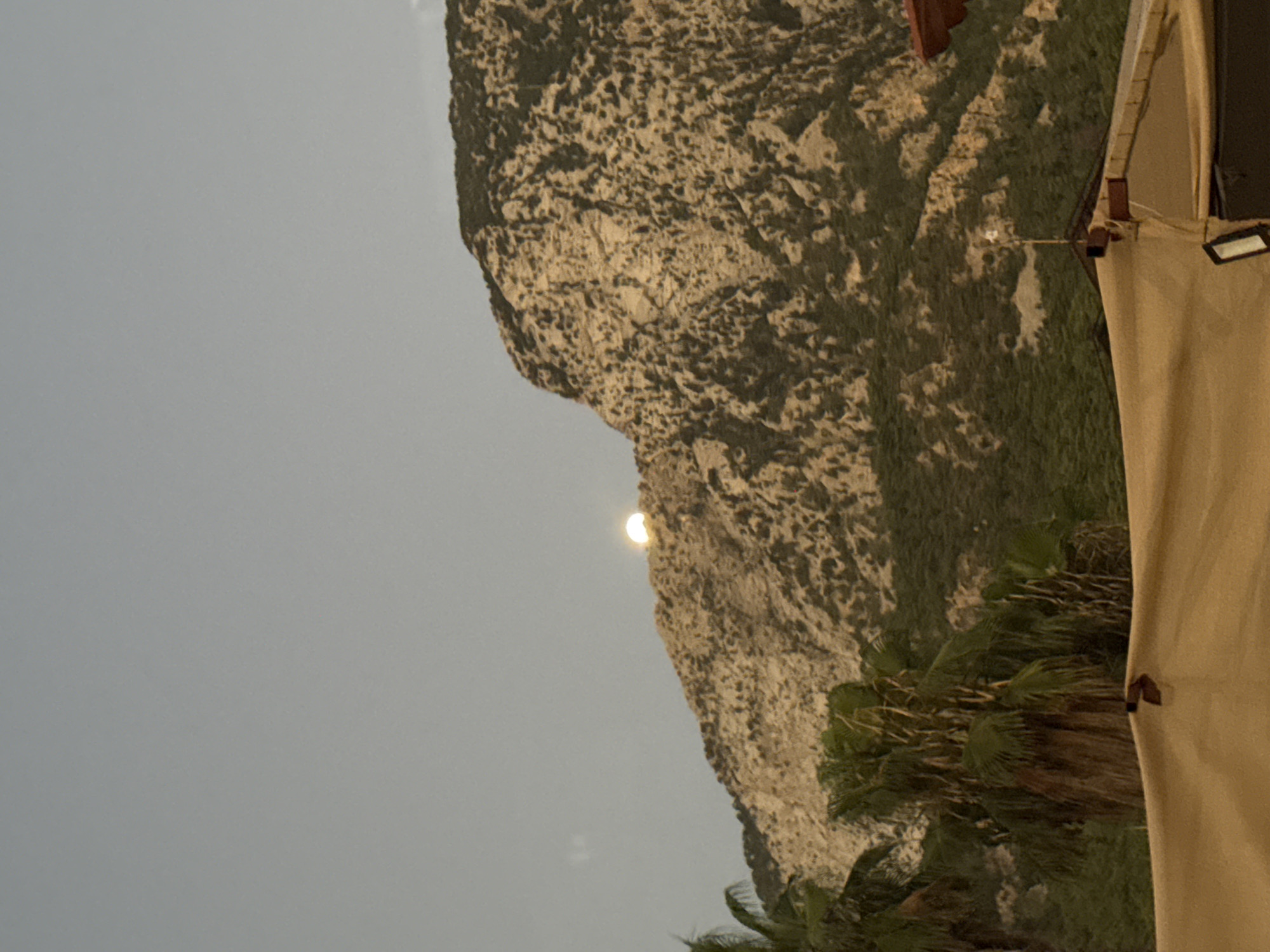 The image shows a mountain landscape with the moon visible behind the summit. Palm trees are in the foreground, with a tan-colored canopy or awning partially covering the bottom of the frame. The sky is a pale, hazy blue. The mountain is rugged and rocky, with patches of dark green vegetation on its slopes. The moon is a bright, circular object, partially obscured by the mountain's peak, suggesting it is either rising or setting. The scene appears to be set in Kemer, Turkey, known for its coastal mountains.