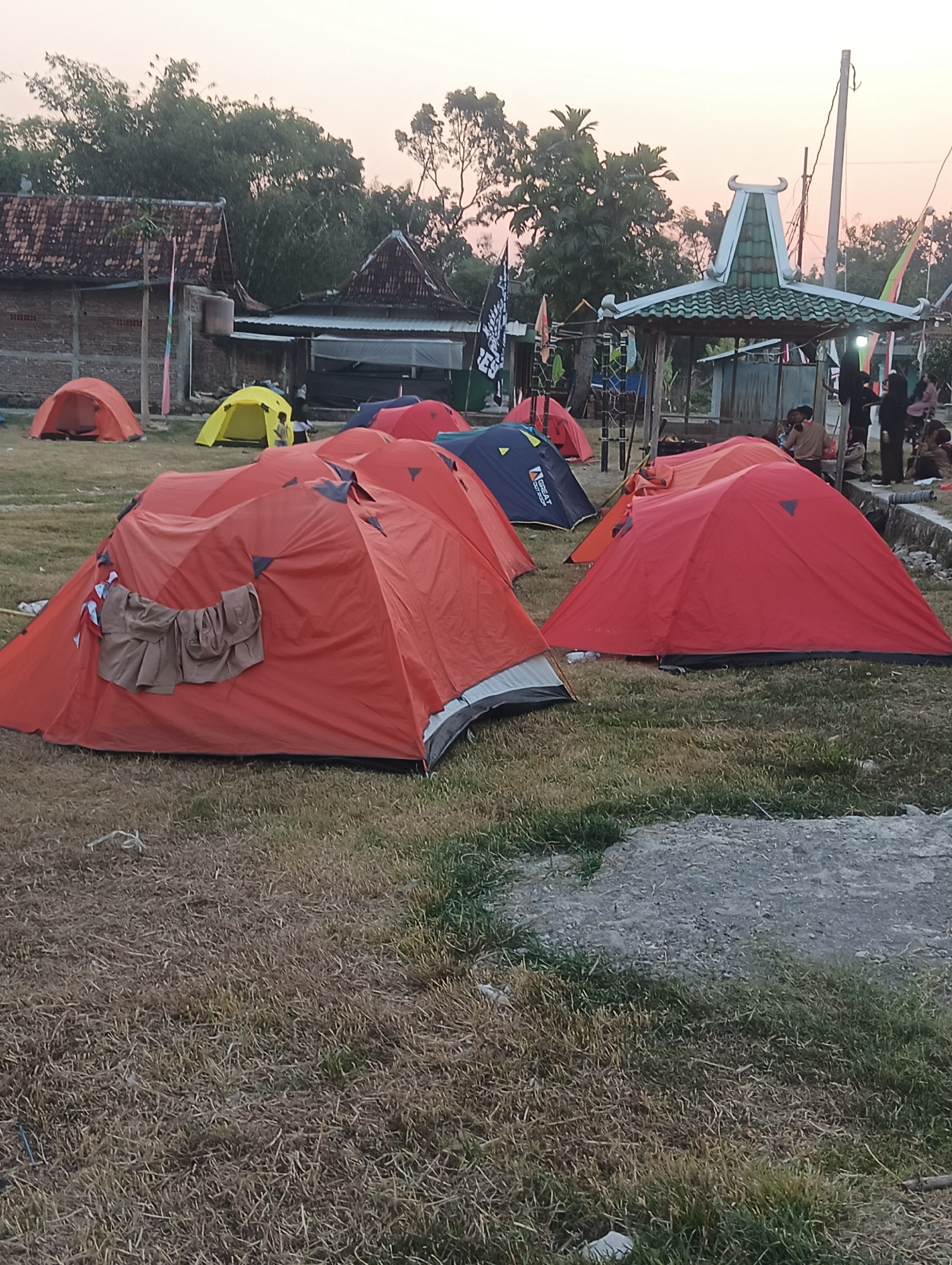 This image depicts an outdoor scene in Pilangsari, Indonesia, featuring a campsite. The foreground is dominated by several camping tents in various colors, primarily red and orange, with one yellow tent further back. Some tents are open, while others are closed. The ground is covered with dry grass and some patches of green grass.
In the background, there are traditional Indonesian-style buildings with tiled roofs. To the right of the frame, a gazebo-like structure with a decorative roof is visible, surrounded by people. Several individuals, dressed in casual attire, are present near the gazebo, some standing and others seated. Various flags are planted around the campsite, including one with black lettering on a white background. The overall lighting suggests it is either early morning or late afternoon, with a soft, diffused sky. The environment appears to be a rural or semi-rural area.