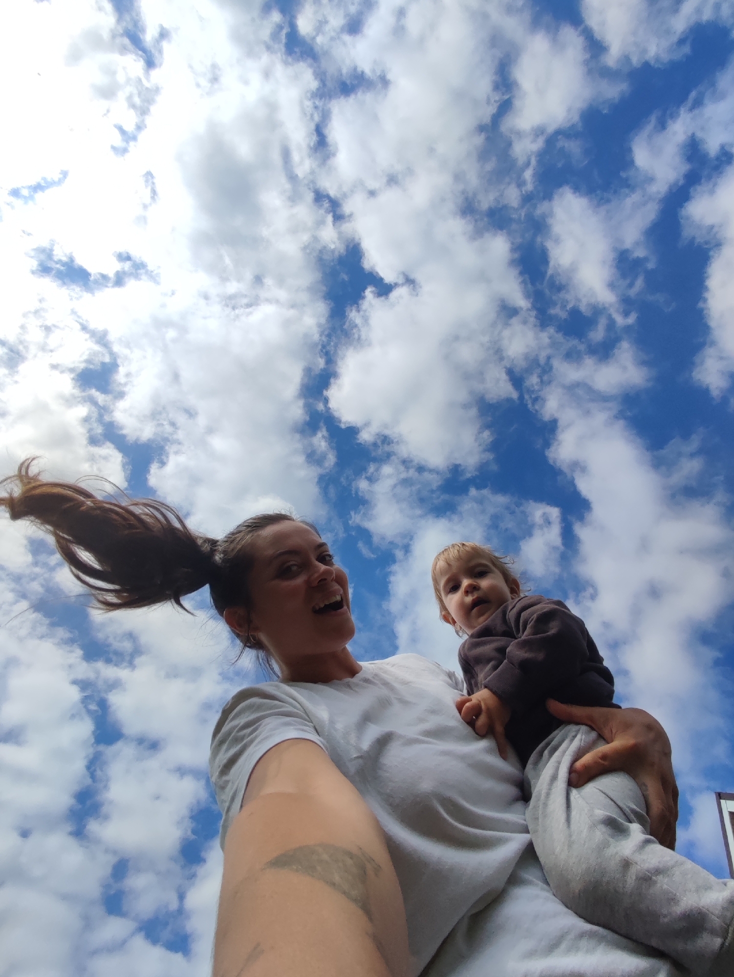 A low-angle, wide shot captures a woman holding a child against a backdrop of a bright blue sky filled with fluffy white clouds. The woman, with her hair pulled back into a ponytail that is blowing to the left, is looking up and to the right with a wide smile and open mouth. She is wearing a white t-shirt and has a tattoo on her left forearm. The child, facing the camera, is being held in the woman's arms, wearing a dark hooded sweatshirt and light grey pants. The image appears to be taken outdoors on a sunny day. The location is specified as Фоки, Russia.