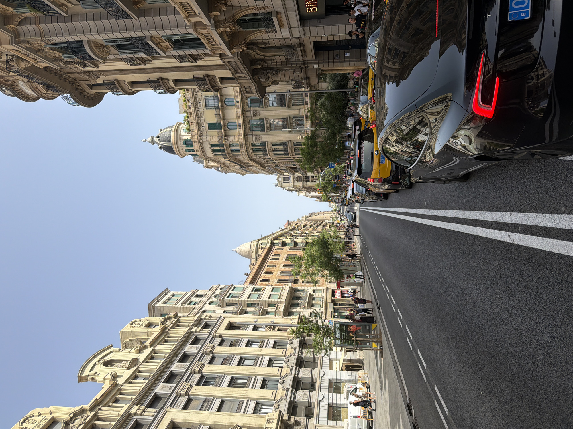 The image captures a bustling street scene in Barcelona, Spain, likely on a sunny day, given the clear blue sky. The perspective is from street level, looking down a wide avenue flanked by ornate, multi-story buildings with neoclassical architecture. These buildings feature detailed facades with balconies, arched windows, and decorative cornices.
On the left side of the street, a pedestrian walkway runs alongside the road. Several people are visible on this walkway, some standing and others walking. A white van is parked on the left, and a bus stop shelter is also present. Trees are planted along the sidewalk.
The road itself is asphalt with clear white lane markings. It is heavily congested with traffic. Several yellow taxis, specifically Freenow branded vehicles, are prominent in the traffic flow, along with other cars. A black electric vehicle, possibly a Tesla, is in the foreground on the right side of the frame, facing away from the camera.
The street is lined with more buildings on the right, similar in architectural style to those on the left. Signs for businesses are visible on the ground floors of these buildings. People are also walking on the sidewalk on the right side of the street. The overall impression is one of urban activity and lively city life.