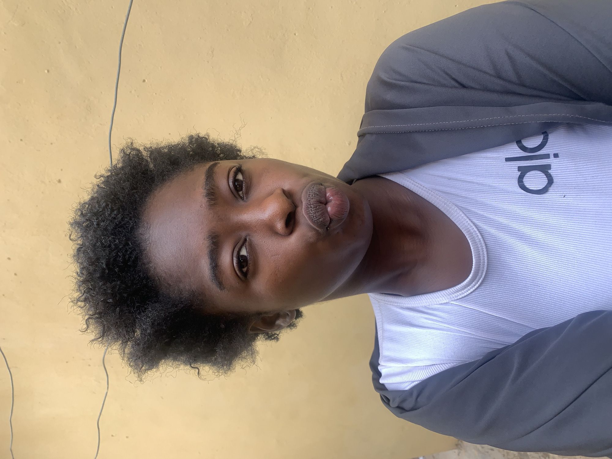 A young Black woman is posing for a selfie. She is wearing a white ribbed tank top with the word "Alo" printed on it and a dark gray jacket. Her hair is short and curly. She is puckering her lips. The background is a yellow wall. The location is most likely in a building in the city of Rafin Par, Nigeria. A thin, possibly electrical wire is visible along the top left of the picture.