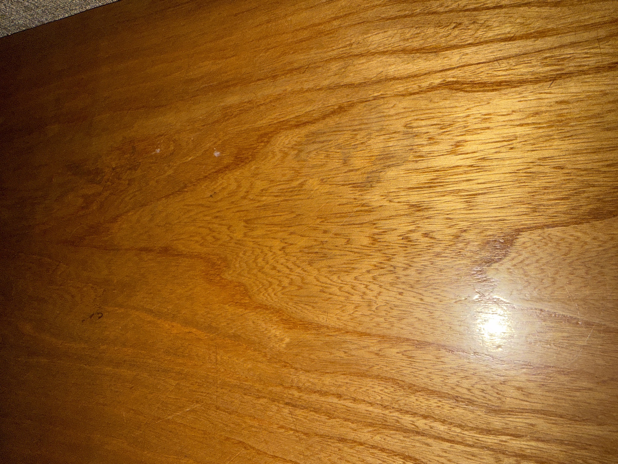 The image shows a close-up view of a wooden surface. The wood grain is prominent, with vertical streaks of darker brown against a lighter brown background. There are some lighter spots and subtle marks on the surface, suggesting it may be a tabletop or a piece of furniture. The lighting creates a bright glare in the lower center of the image. The top right corner of the image shows a small portion of a grey, textured material, possibly fabric or a carpet. There are no people, animals, or any specific landmarks visible that would indicate a location in Okhtyrka, Ukraine. The image is a detail shot focused solely on the texture and appearance of the wood.