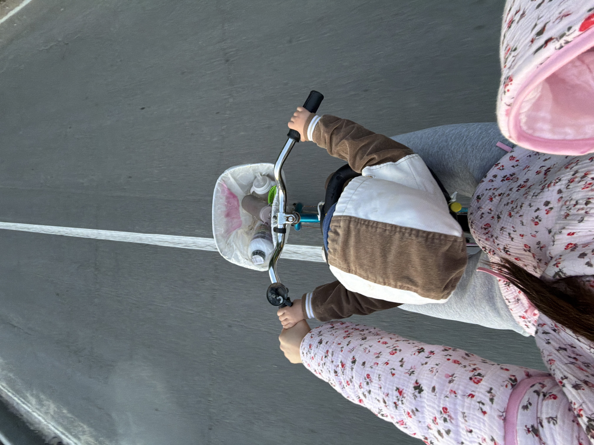This image shows a close-up, overhead view of two people on a bicycle, seemingly moving on a paved surface. The foreground features the arm and shoulder of an adult wearing a light pink, floral-patterned garment and a pink hood. Their arm is visible across the handlebars, and their hand appears to be either holding on or resting near the child's hand.
The middle ground is dominated by a child on the bicycle, wearing a brown and white jacket with white stripes on the sleeves. The child's hands are on the handlebars, with one hand gripping the right handlebar. A white basket is attached to the front of the bicycle, containing a water bottle. The bicycle itself has a blue frame.
The background is a gray, paved surface, likely a road, with a white line running horizontally across it. The lighting suggests it is daytime, possibly late afternoon given the angle of the sun casting subtle shadows. The overall impression is one of a casual outing or a journey, possibly in a park or on a quiet street, given the focus on the people and the bicycle. The image is cropped tightly, focusing on the interaction and the movement of the bicycle. There is no visible text in the image. The setting appears to be an outdoor environment with a paved pathway. The emotional tone is one of shared activity and perhaps a sense of gentle movement and companionship.