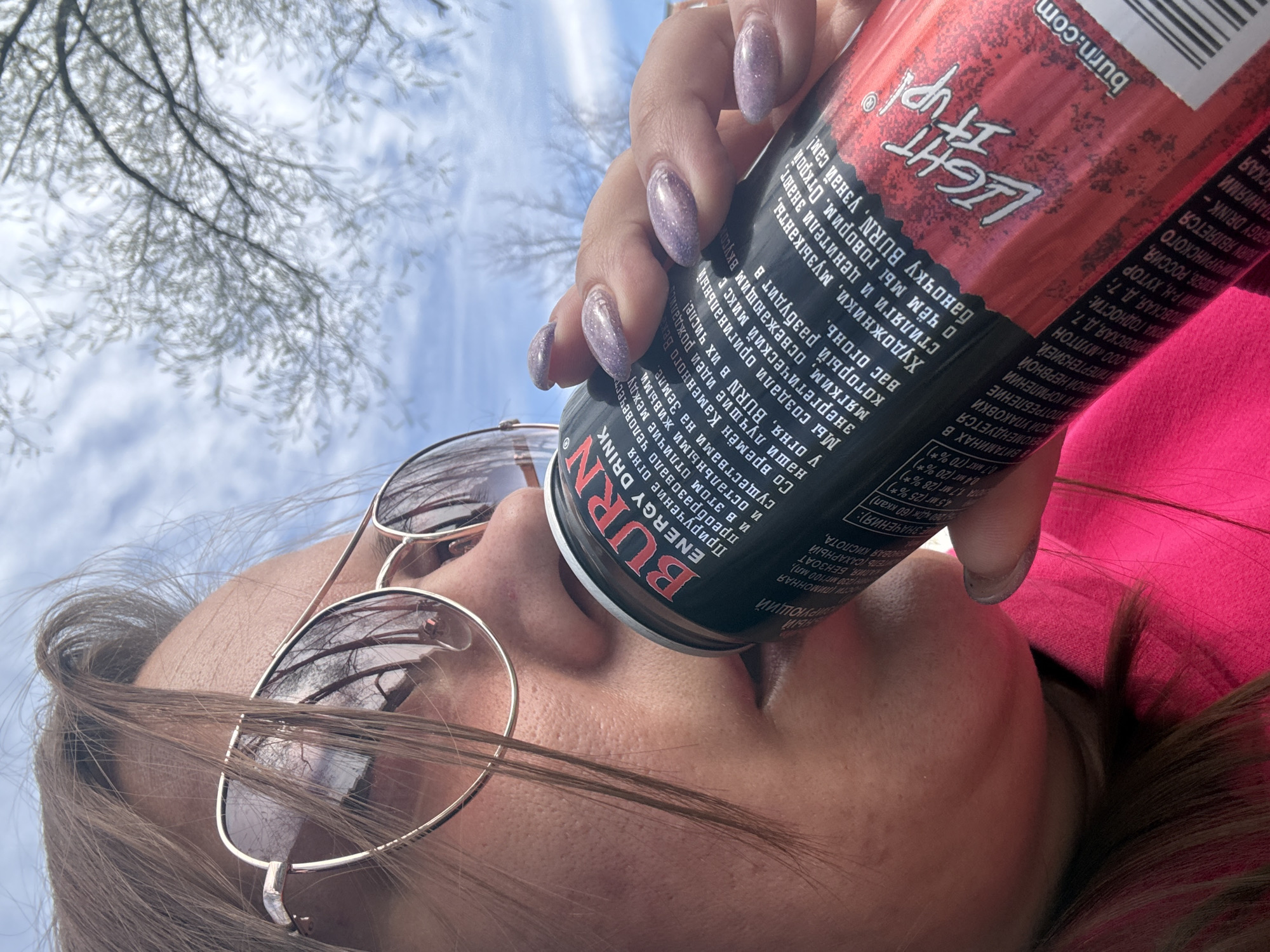 A woman is drinking from a can of BURN Light Up energy drink. She is wearing sunglasses and has light purple nail polish. The can of BURN energy drink has text in Russian on it. The barcode indicates that the drink can be purchased in Dmitrov, Russia. The background shows a blue sky and tree branches. The woman is wearing a pink shirt or jacket.