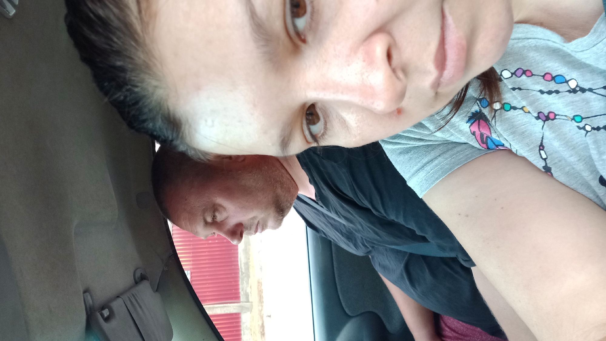 Two individuals, a man and a woman, are seated inside a vehicle. The man is in the driver's seat, looking to his left. The woman is in the passenger seat, looking forward and slightly towards the camera. The interior of the car is visible, including the dashboard and a sun visor. Outside the car, a red fence and some vegetation can be seen, suggesting an urban or suburban environment. The image was taken in Kropyvnytskyi, Ukraine.