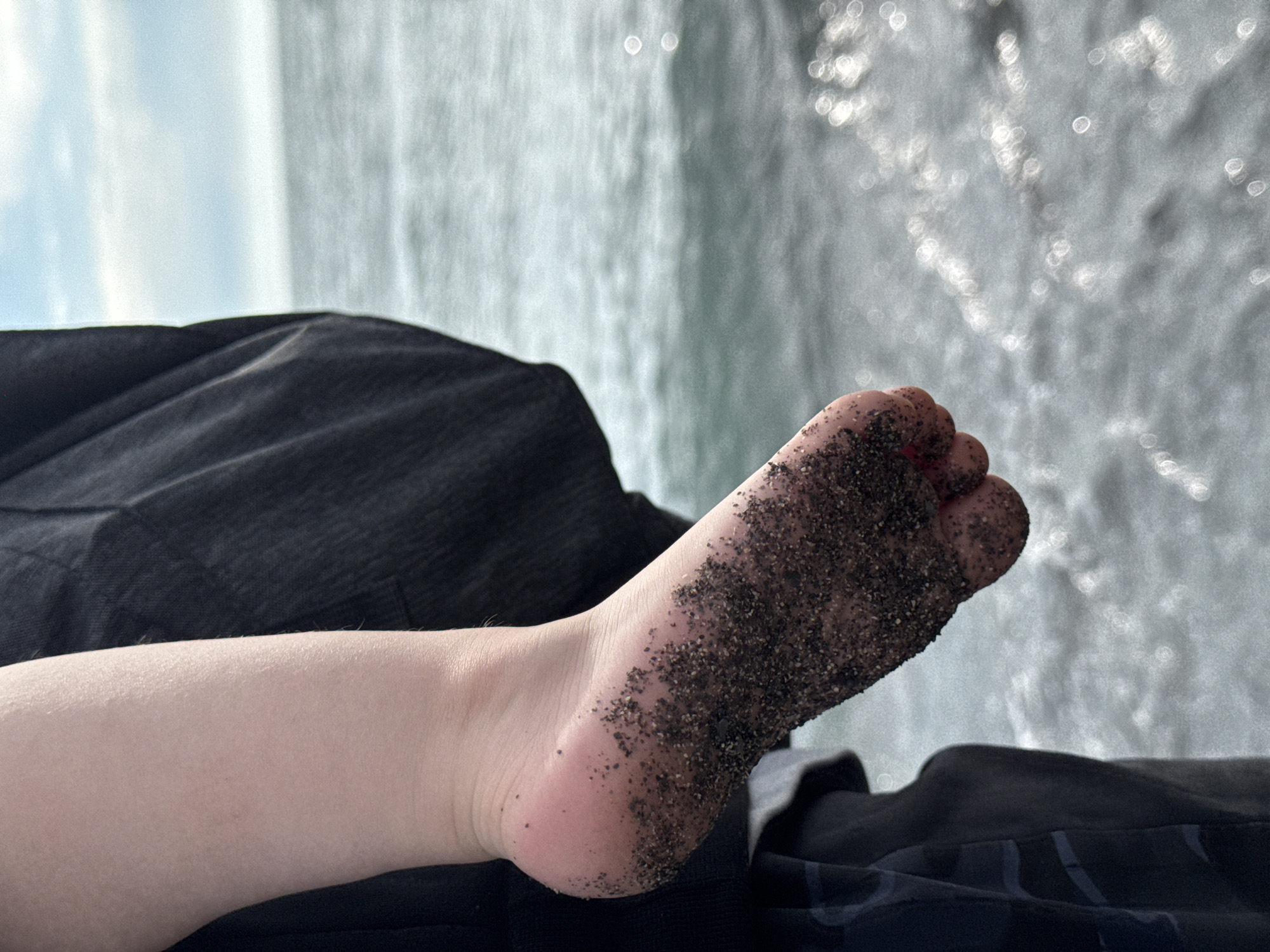 A close-up photograph shows a person's bare foot covered in dark sand. The foot is extended, with the toes pointed upwards. The skin of the foot is light-toned. The person is wearing dark clothing, some of which is visible in the background. The backdrop appears to be a body of water with blurred, bokeh-like reflections, which might indicate a sunny day. Given the location in Sochi, Russia, the body of water is likely the Black Sea.