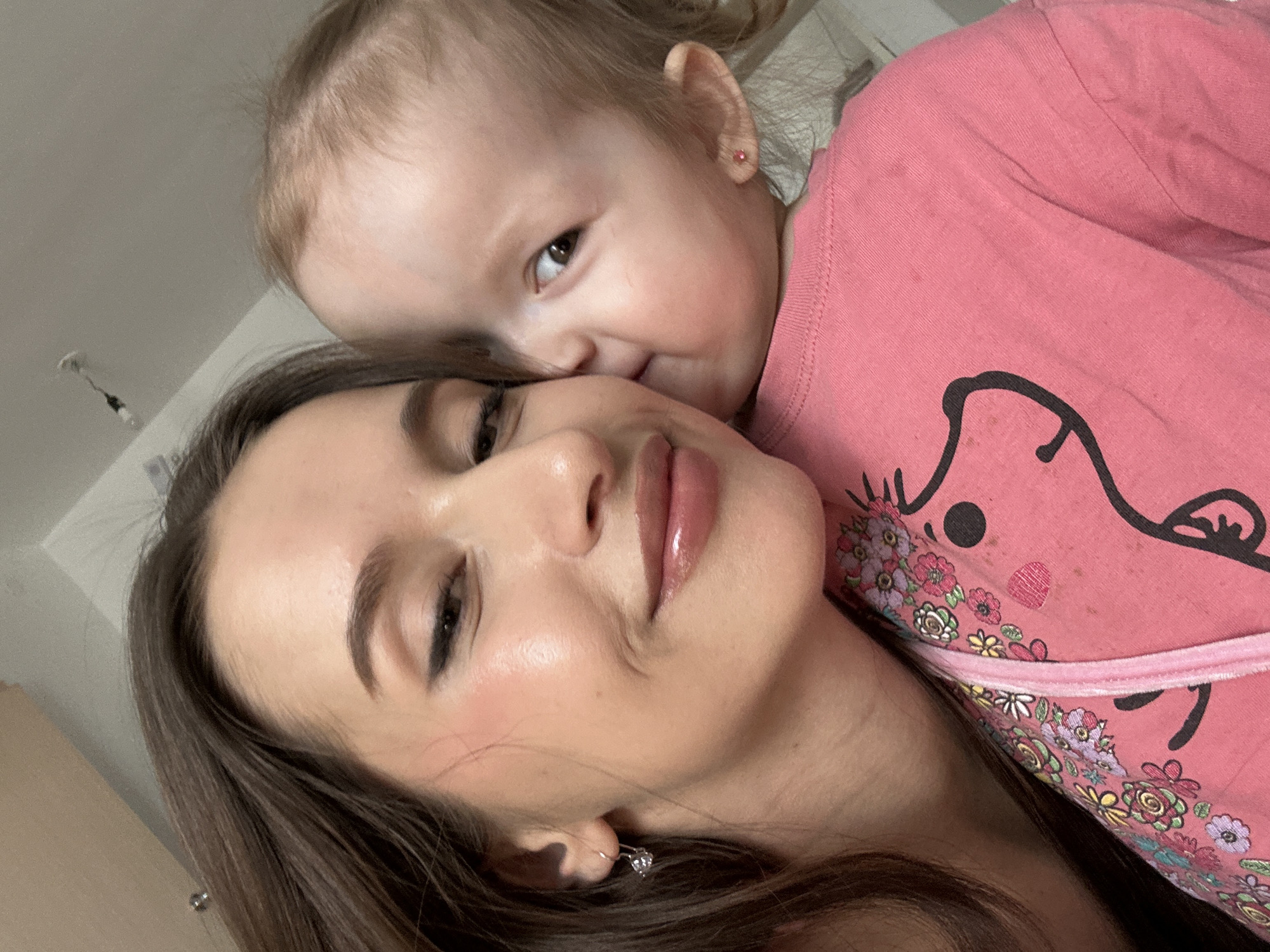A close-up, selfie-style photograph shows a young woman with long brown hair and a baby, likely her child, snuggling close to her face. The woman has fair skin, brown eyes, and full lips, with what appears to be glossy makeup. She is wearing earrings. The baby, who has fair skin and light brown hair, is wearing a pink shirt with a graphic design. The background is a plain light-colored wall. The scene is likely indoors. There is no information available to determine if the image was taken in Yekaterinburg, Russia.