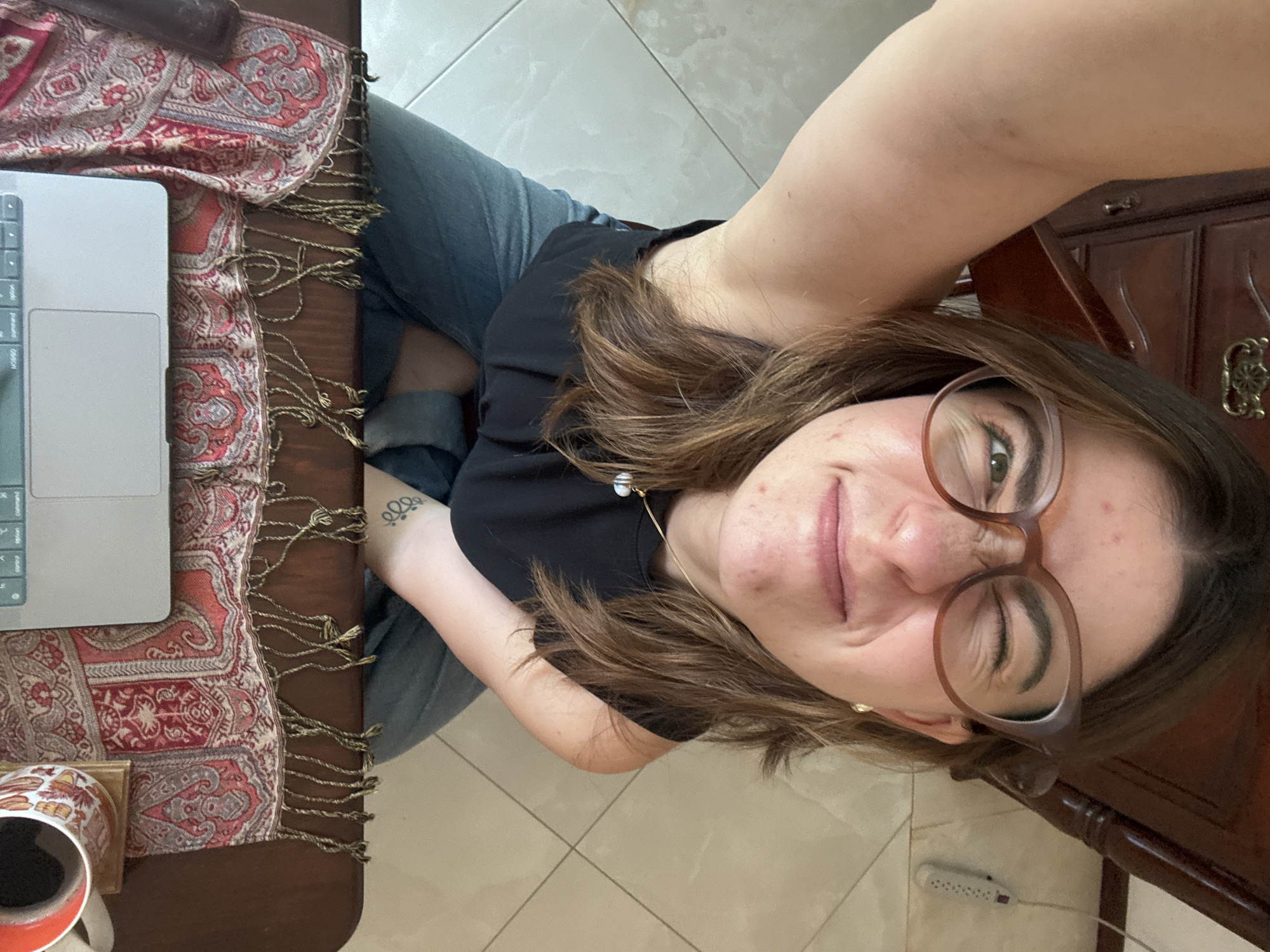 A woman with brown hair and glasses is seated at a wooden table covered with a red patterned scarf. She is wearing a black shirt and jeans. A laptop and a mug are on the table to her left. The background shows a tiled floor and a dark wooden cabinet. A power strip is visible on the floor near the cabinet. The location is likely an indoor setting in Puebla, Mexico.