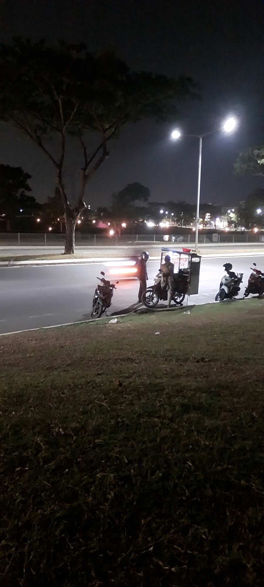 In the city of Medan, Indonesia, at night, a street scene unfolds. A large tree stands to the left of the frame, its branches silhouetted against the dark sky. A tall lamppost with two bright lights illuminates the area. In the foreground, a grassy embankment slopes down to a paved road.
On the road, several motorcycles are parked or in motion. Two individuals are visible near a motorcycle with a sidecar, which appears to be a food cart with a sign on it. One person is standing, wearing a helmet, and holding onto the handlebars. Another person is seated on the motorcycle. Further down the road, two more motorcycles are present, one with a rider. The scene is lit by streetlights and the headlights and taillights of the vehicles, creating streaks of light. The overall atmosphere is that of a quiet night on a city street, possibly a roadside stall or a temporary stop.