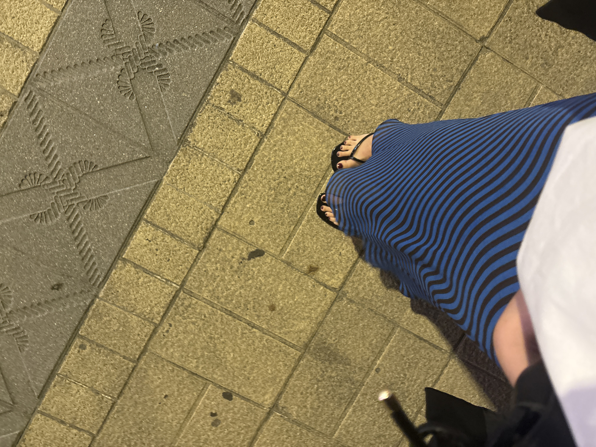 The image shows a person's feet wearing black flip-flops and a long blue and black striped dress. The person is standing on a sidewalk made of tan and gray paving stones. The paving stones have a decorative pattern on some of the gray sections. The scene appears to be outdoors, possibly in a city. The lighting suggests it might be evening or nighttime, as indicated by the shadows and the yellow cast on the paving stones. There is also a hint of a black object, possibly a bag or umbrella, to the left of the frame. The location is identified as Sanxenxo, Spain.