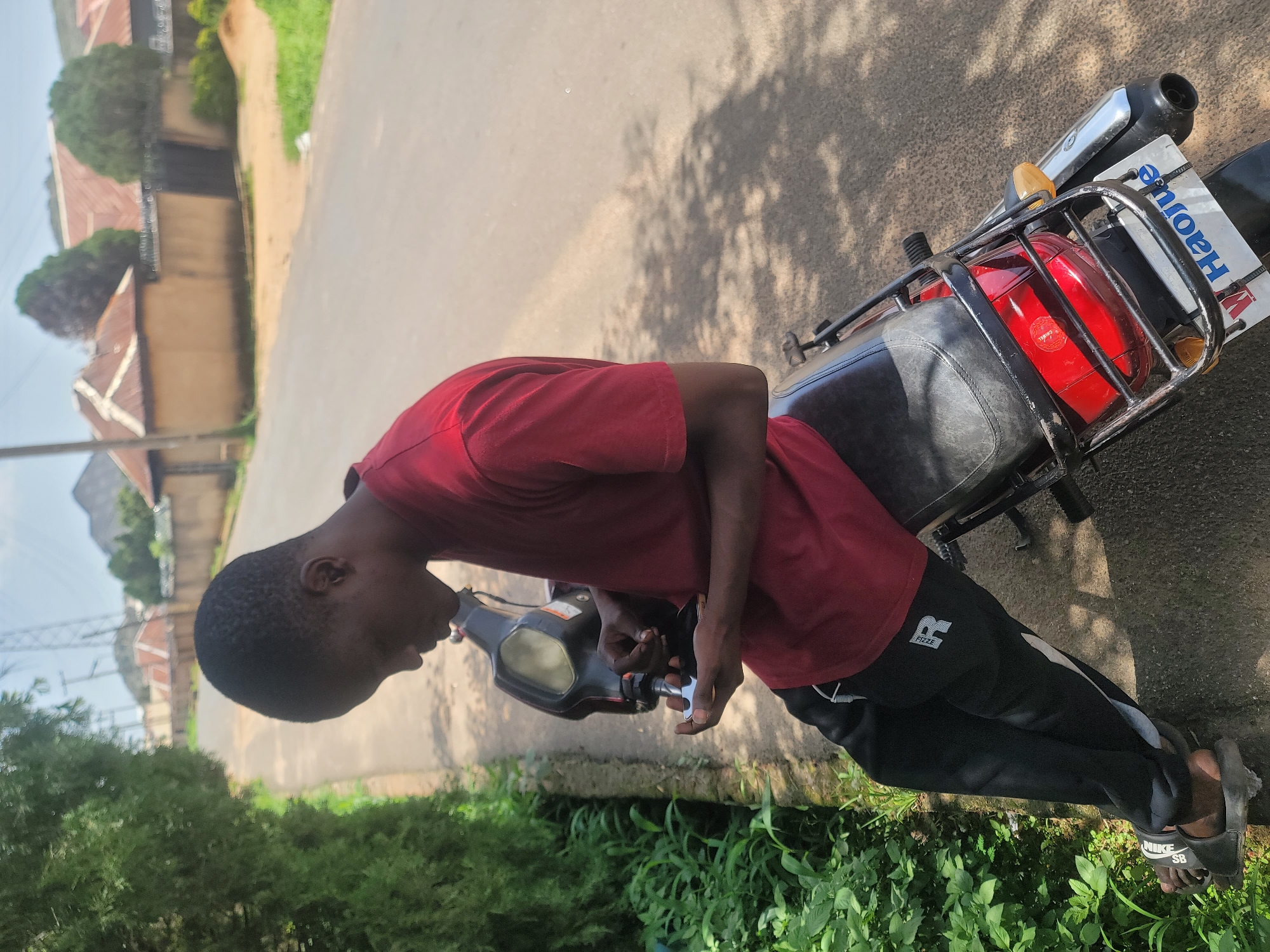 A young Black male is seated on a parked motorcycle on a paved road in Jos, Nigeria. He is wearing a red t-shirt and black sweatpants with an "R" logo on the left thigh. The motorcycle is black with a red taillight and a license plate that reads "Haojue". The scene is outdoors with lush green foliage on the left side of the frame, a paved road, and houses in the background. The sky is visible with some clouds. The man appears to be looking down at something in his hands, possibly adjusting a part of the motorcycle or his phone.