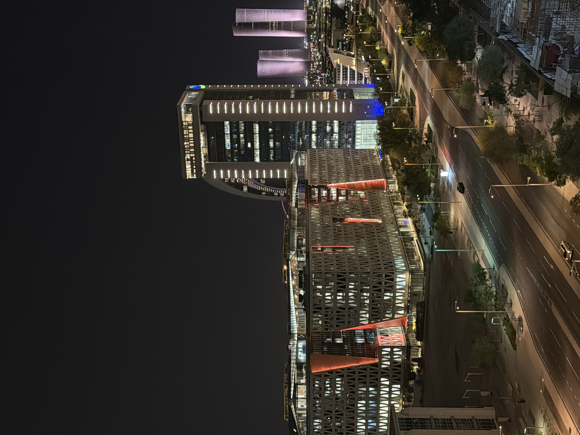 A nighttime, elevated, wide-angle view shows a modern city street with contemporary buildings on the left and background. The sky is dark, indicating night. In the foreground, a paved road with multiple lanes curves to the right, bordered by sidewalks and streetlights. Several cars are visible on the road, with headlights and taillights illuminated. Trees and landscaping line the street.
Dominating the left side of the frame are several buildings. The closest building is a low-rise structure with a complex, geometric facade featuring a patterned exterior and illuminated red triangular accents. Behind this, a tall, modern skyscraper with a curved facade is visible, featuring vertical white lighting. Further in the background, other tall skyscrapers rise into the night sky, some with purplish illumination. The buildings are lit from within, showcasing their interiors. The overall scene depicts a well-developed urban environment at night.