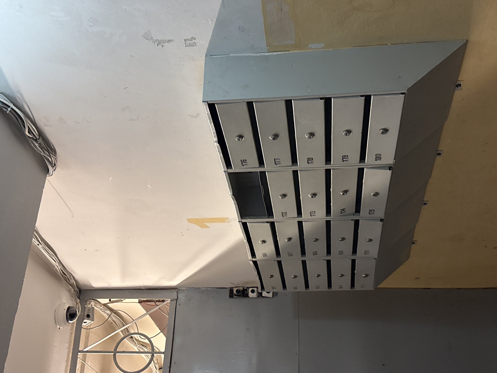 The image captures a cluster of silver mailboxes, numbered from 165 to 180, mounted on a wall. The top row of boxes is numbered 176 through 180. The mailboxes have small keyholes and are evenly spaced. The wall on which they are mounted appears to be painted in light gray. Above the mailboxes, there is a section of the wall painted in a yellow-beige hue. The ceiling is also painted in a light gray color, with some discolored patches and exposed wiring running along the corner. In the lower-left corner of the image, there is a glimpse of a surveillance camera mounted on the wall, and a partially visible doorway or opening with a metal security grill.
While the location of Izhevsk, Russia is based on general knowledge that apartment buildings often have a collection of mailboxes. However, there is no definitive information provided within the image itself to confirm the location.