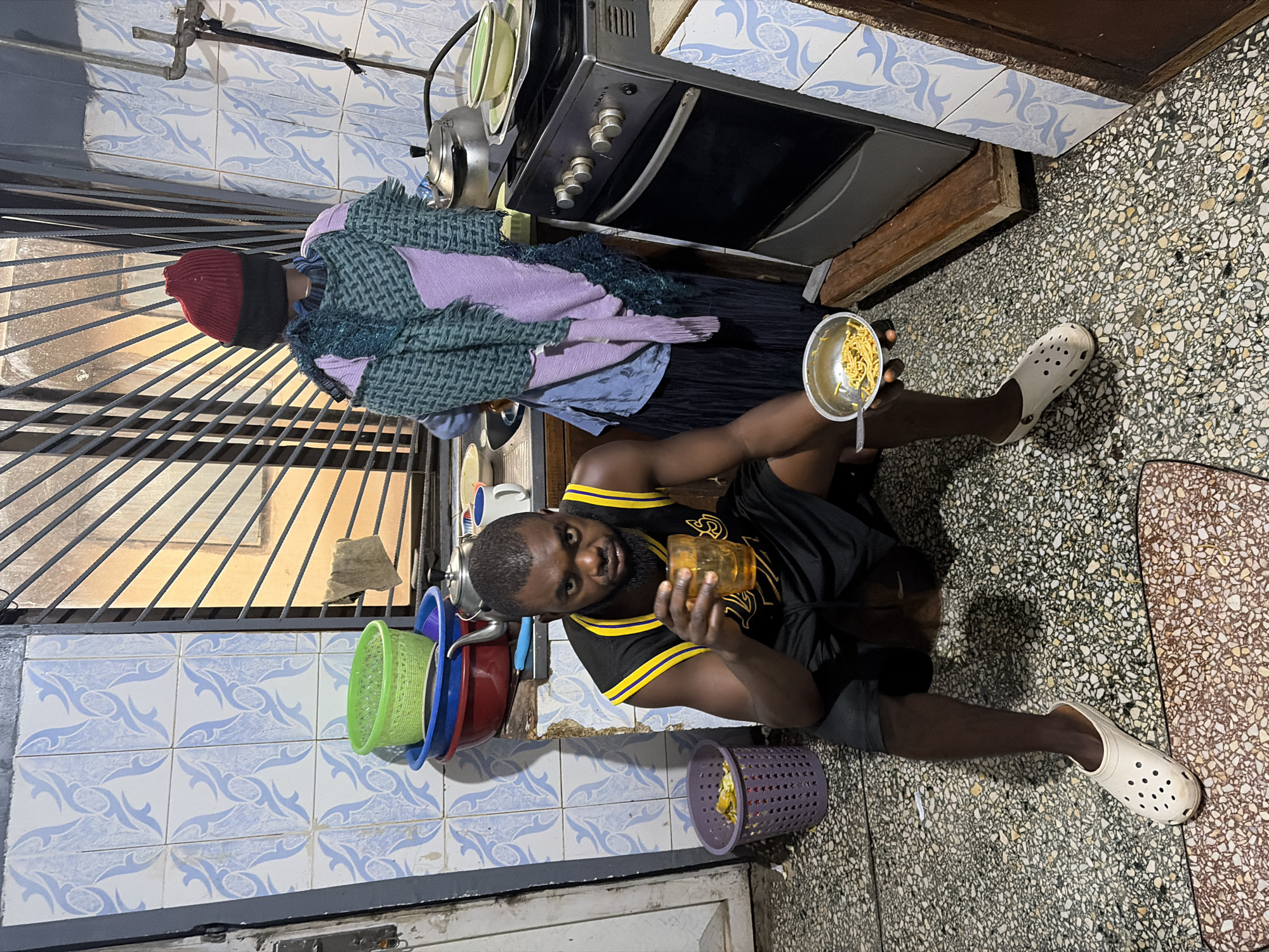 A young Black man is seated on the floor in a dimly lit, cramped kitchen, likely in Jos, Nigeria. He is wearing a black basketball jersey with yellow trim and black basketball shorts, along with white Crocs. He holds a glass of amber liquid in his right hand and a small metal bowl of noodles in his left, with a fork resting in the bowl. To his left is a purple plastic basket containing what appears to be fruit peels. Behind him, a stove is visible. To the left of the stove, a mannequin is dressed in a blue shirt and a purple and green patterned wrap, topped with a red beanie. The kitchen walls are tiled with a blue and white pattern, and a barred window is behind the stove and mannequin. The floor is covered in a speckled terrazzo material.