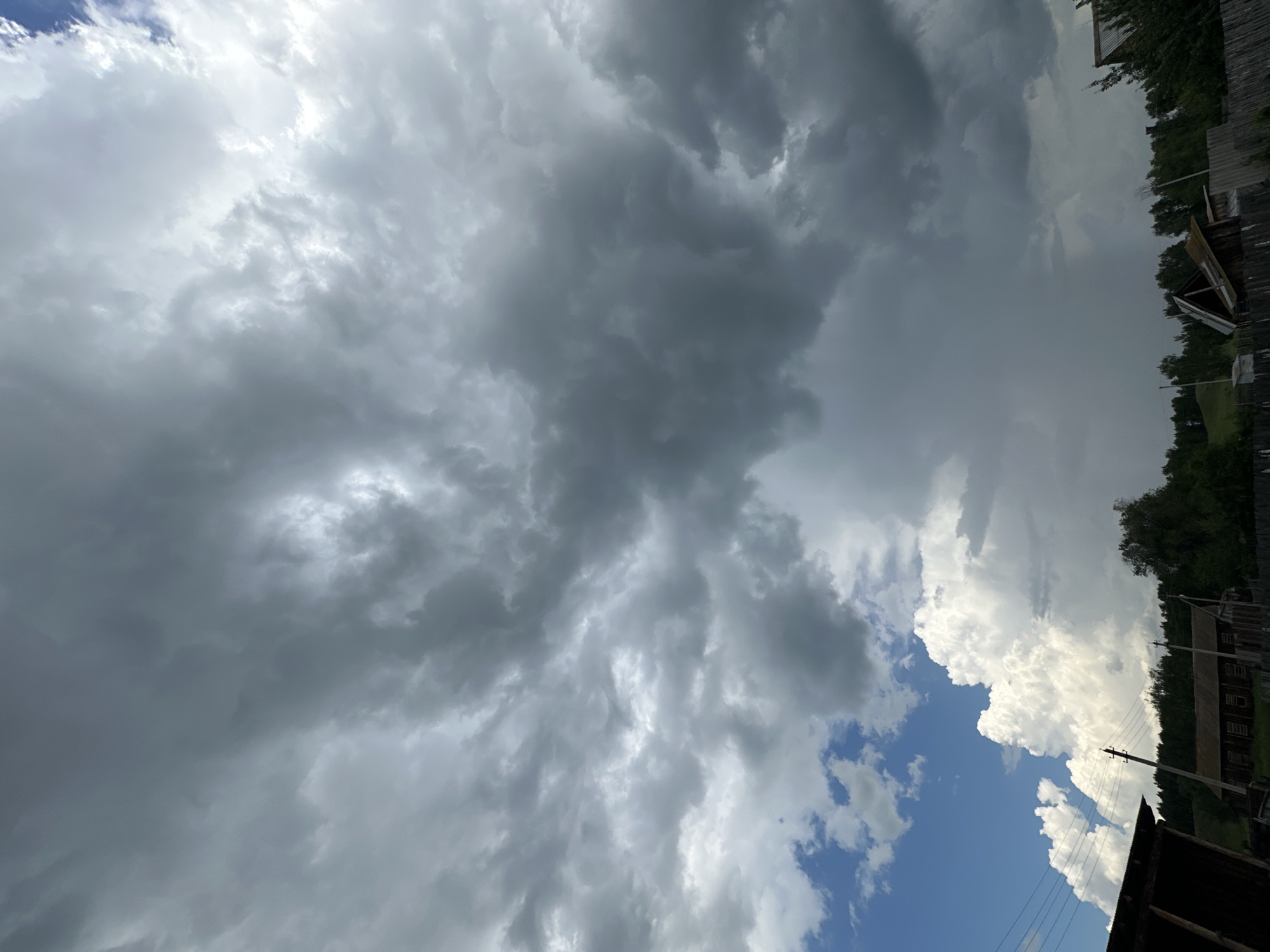The image shows a dramatic sky filled with dark, heavy storm clouds. In the lower portion of the image, a small village is visible, with wooden houses and trees. The sky is a mix of dark grays and blues, with some bright white clouds interspersed. The overall impression is one of an impending storm.