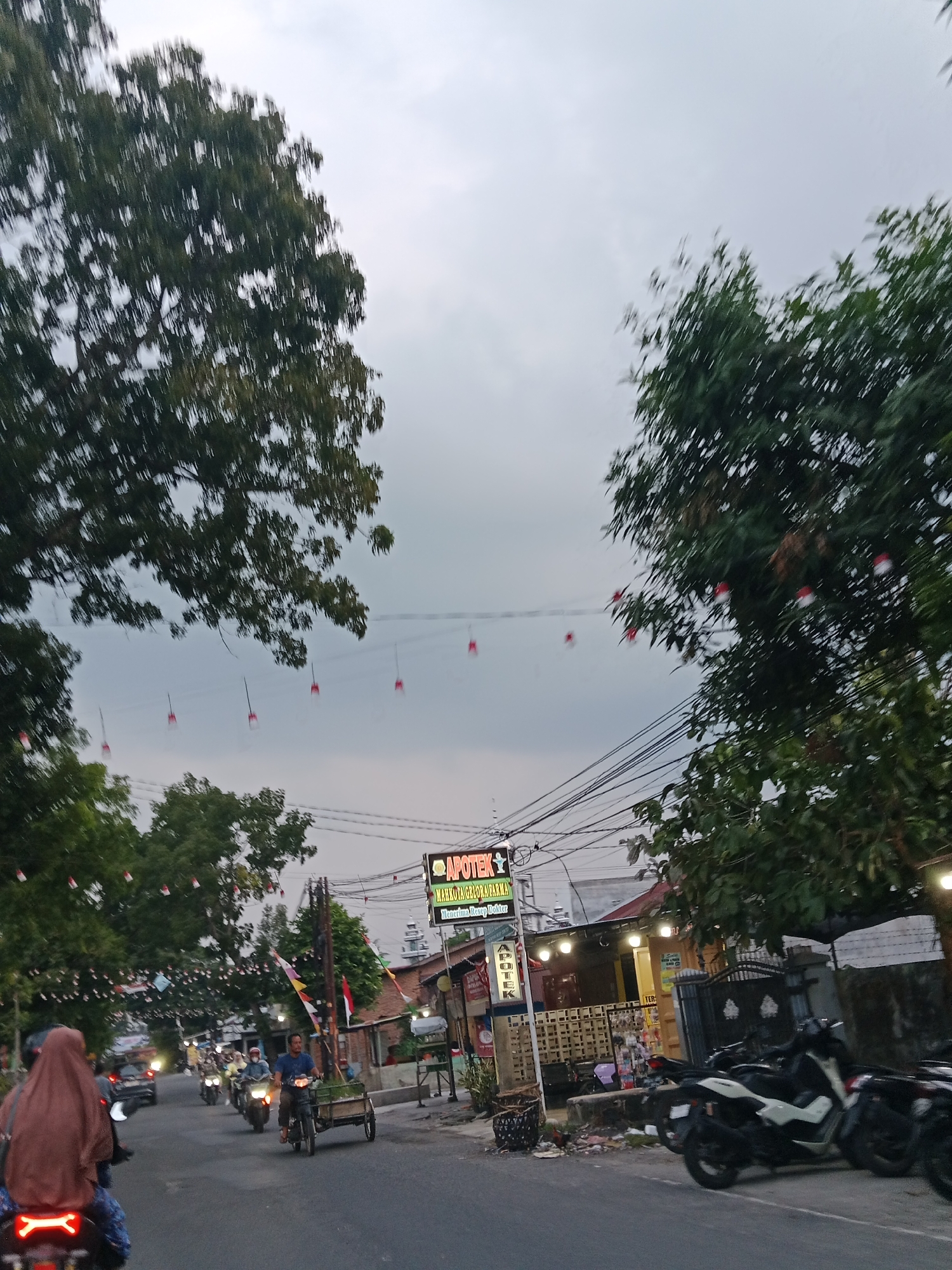 The image depicts a street scene in the city of Binjai, Indonesia, likely during dusk. A road runs through the center, with buildings lining the right side. Several motorcycles and a bicycle with a cart are visible on the road, suggesting moderate traffic. A few individuals are on the motorcycles, with one person in the foreground riding a motorcycle and wearing a brown hijab.
The buildings on the right appear to be shops or commercial establishments, with one prominently featuring a sign for "APOTEK" (pharmacy). Decorative lights or banners, some appearing to be Indonesian flags, are strung across the street and along the trees. The sky is overcast, contributing to the subdued lighting. The environment is urban, with trees, power lines, and paved roads.