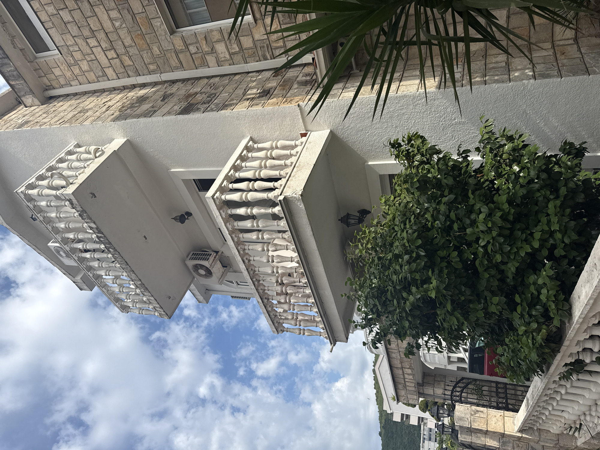 An eye-level shot, rotated ninety degrees counter-clockwise, displays the exterior of a multi-story building in Budva, Montenegro, under a bright, sunny daytime sky with scattered white clouds. The building features light-colored, likely white or cream-colored, stucco walls with sections of decorative, light-brown stone tiling on its upper facade.
Multiple balconies are visible, each adorned with prominent, ornate white balustrades composed of many small balusters. Two air conditioning units are mounted on the exterior walls, one clearly seen on the leftmost balcony and another partially visible on a higher section. Lush green foliage from a large, dense bush fills the lower right portion of the frame, partially obscuring the building's ground level. From the top of the frame, sharp, pointed fronds of a palm tree extend downwards, casting subtle shadows on the building.
The architectural style, characterized by the stucco, balustrades, and presence of AC units, suggests a Mediterranean or similar warm climate. The clear blue sky and strong natural light indicate it is daytime. A distant view of a mountain is faintly visible on the horizon in the lower left of the original image, confirming the geographical context of Budva, Montenegro. No people are visible, and there is no discernible text. The scene is static, capturing architectural and natural elements.