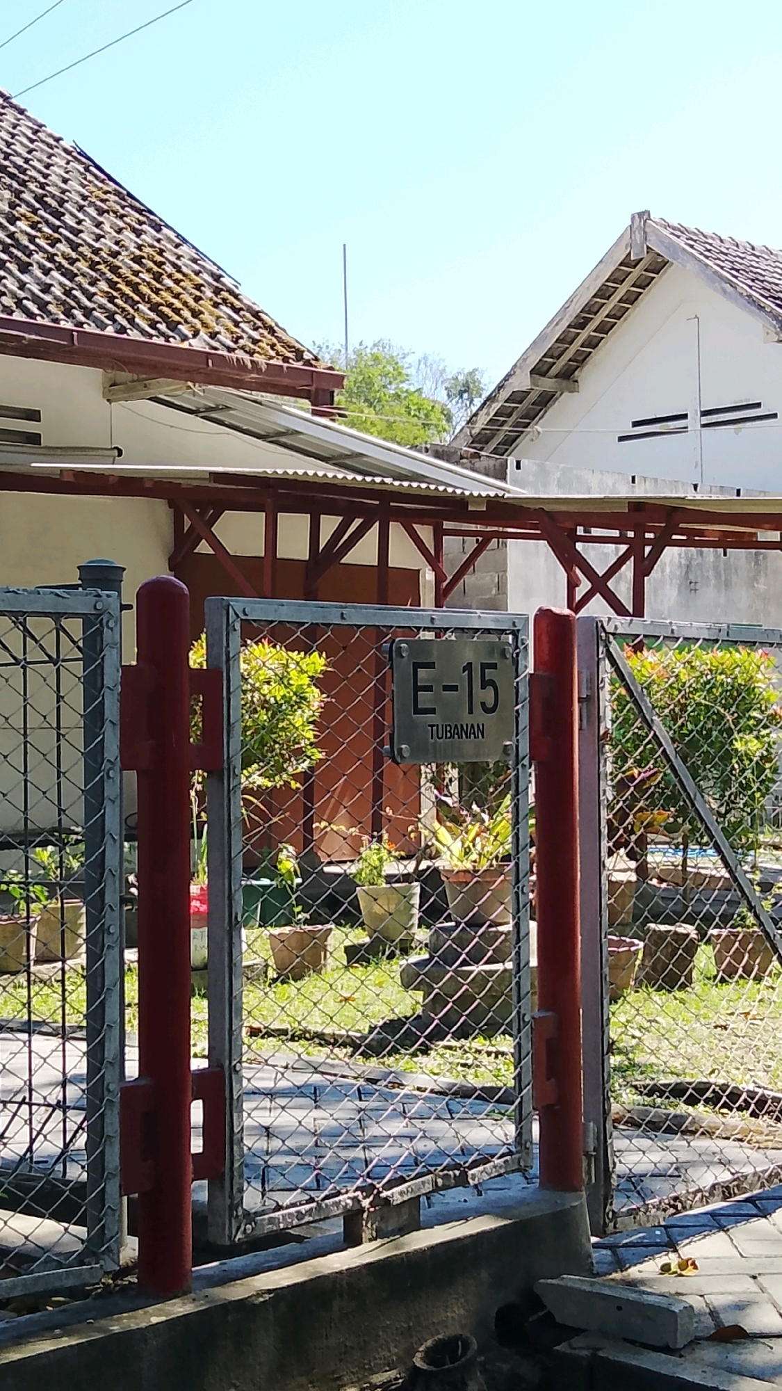 A rectangular metal plaque bearing the inscription "E-15 TUBANAN" is affixed to a chain-link fence in front of a residential property in Tlogobendung, Indonesia. The plaque is positioned at eye level and is visible through the diamond-shaped openings of the fence. Behind the fence, a well-maintained yard is visible, featuring several potted plants. The property has a tiled roof, typical of Indonesian architecture, and a covered patio area. The scene is bathed in bright sunlight, casting distinct shadows on the ground. The overall impression is one of a peaceful, well-kept urban environment.