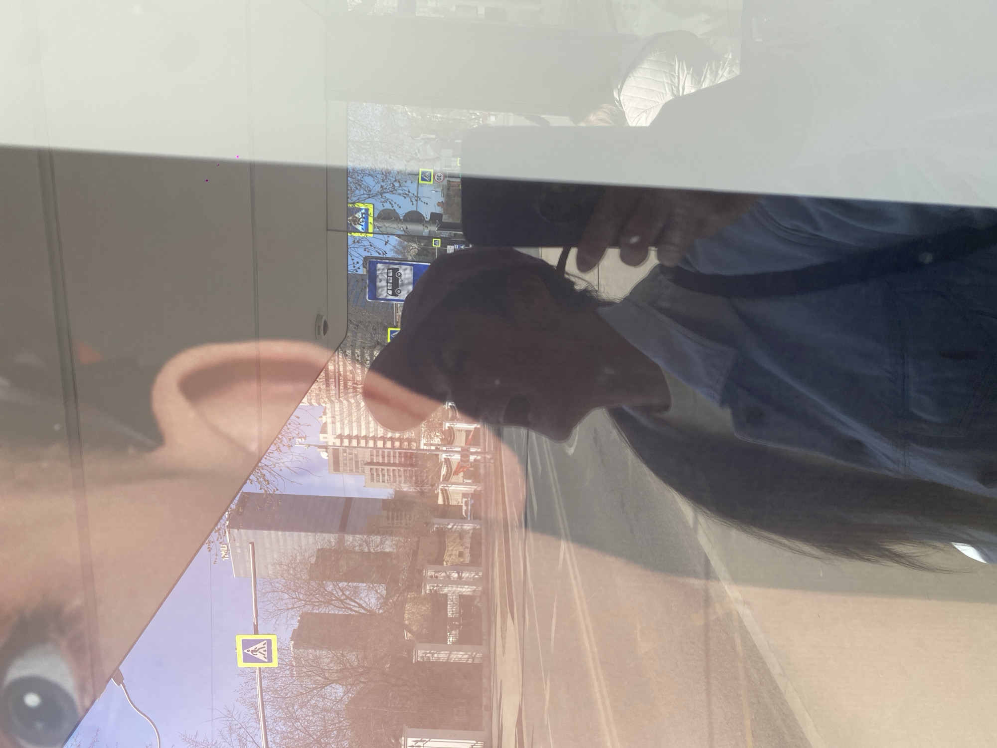 The image shows a reflection of a person holding a mobile phone, seemingly taking a picture of the city outside a window, with the buildings and signs suggesting a location in Moscow, Russia. A pedestrian crossing sign, along with city buildings and infrastructure, are visible in the reflection. The person in the reflection is wearing a light blue shirt. Their reflection is also overlaid on their face.
