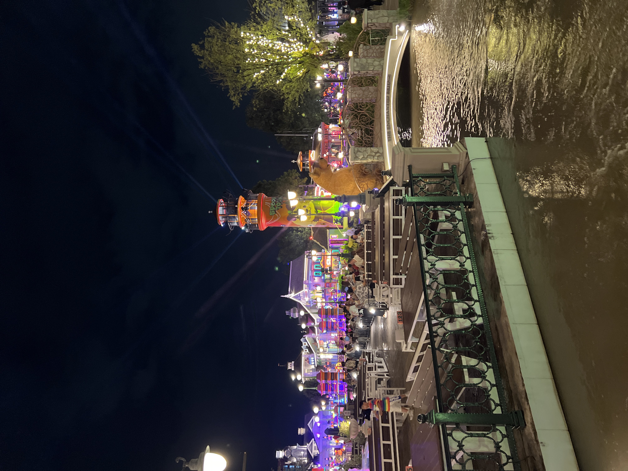 This image depicts a lively nighttime scene at a themed attraction in Bangkok, Thailand. The environment is characterized by a canal or river flowing through the center, spanned by an arched bridge labeled "THE ENCHANTED BRIDGE". The area is densely populated with people dining at numerous outdoor tables and benches, illuminated by a vibrant display of colorful lights on the surrounding buildings and structures. Prominently featured is a tall, multi-colored tower resembling a lighthouse, adorned with graphic elements. Adjacent to the bridge, a large, fuzzy brown bear statue sits. The water reflects the ambient lights, creating a shimmering effect. Trees in the background are also decorated with lights. The overall atmosphere is festive and bustling, suggesting a popular entertainment venue or dining area.